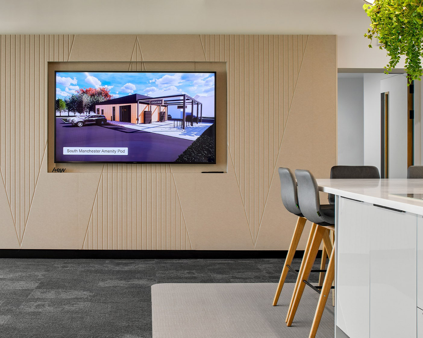 A modern conference room features sleek wooden accent walls complemented by acoustic felt wall panels. A large screen displays an architectural rendering of the "South Manchester Amenity Pool." A white table with high chairs and hanging plants add to the contemporary decor.