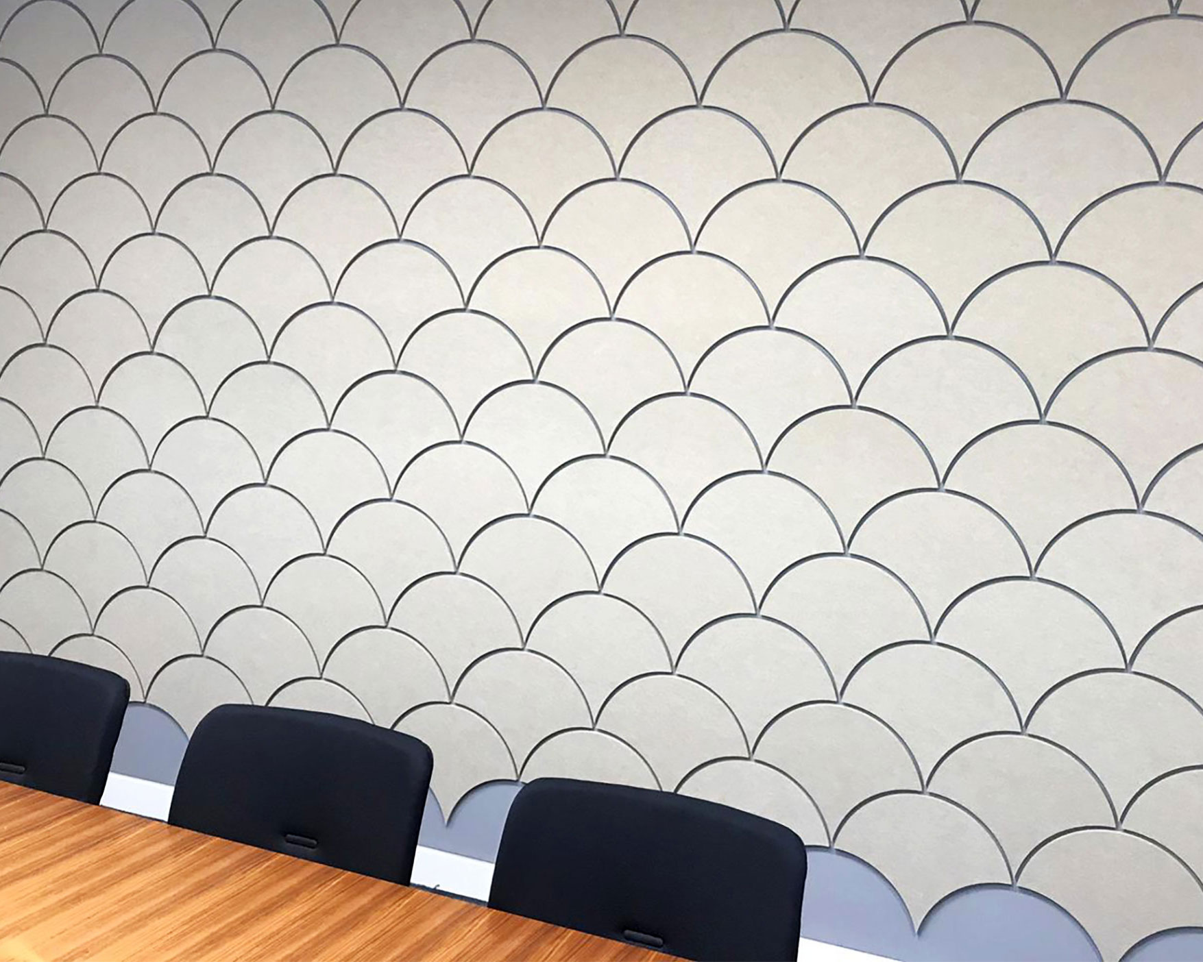 A close-up of an office conference room wall decorated with a scale pattern design in neutral tones using acoustic felt wall tiles. The room features a wooden table and black chairs positioned against the wall. The design creates a modern, stylish ambiance.