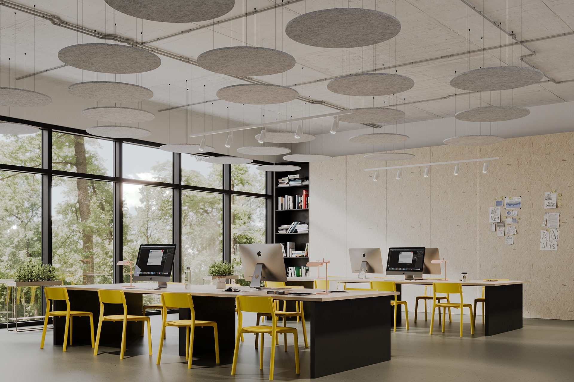 A modern, open-concept office space featuring long shared desks with desktop computers and yellow chairs. Large windows line the wall, offering a view of trees outside. Circle acoustic felt ceiling clouds are suspended from the ceiling, and bookshelves line the back.