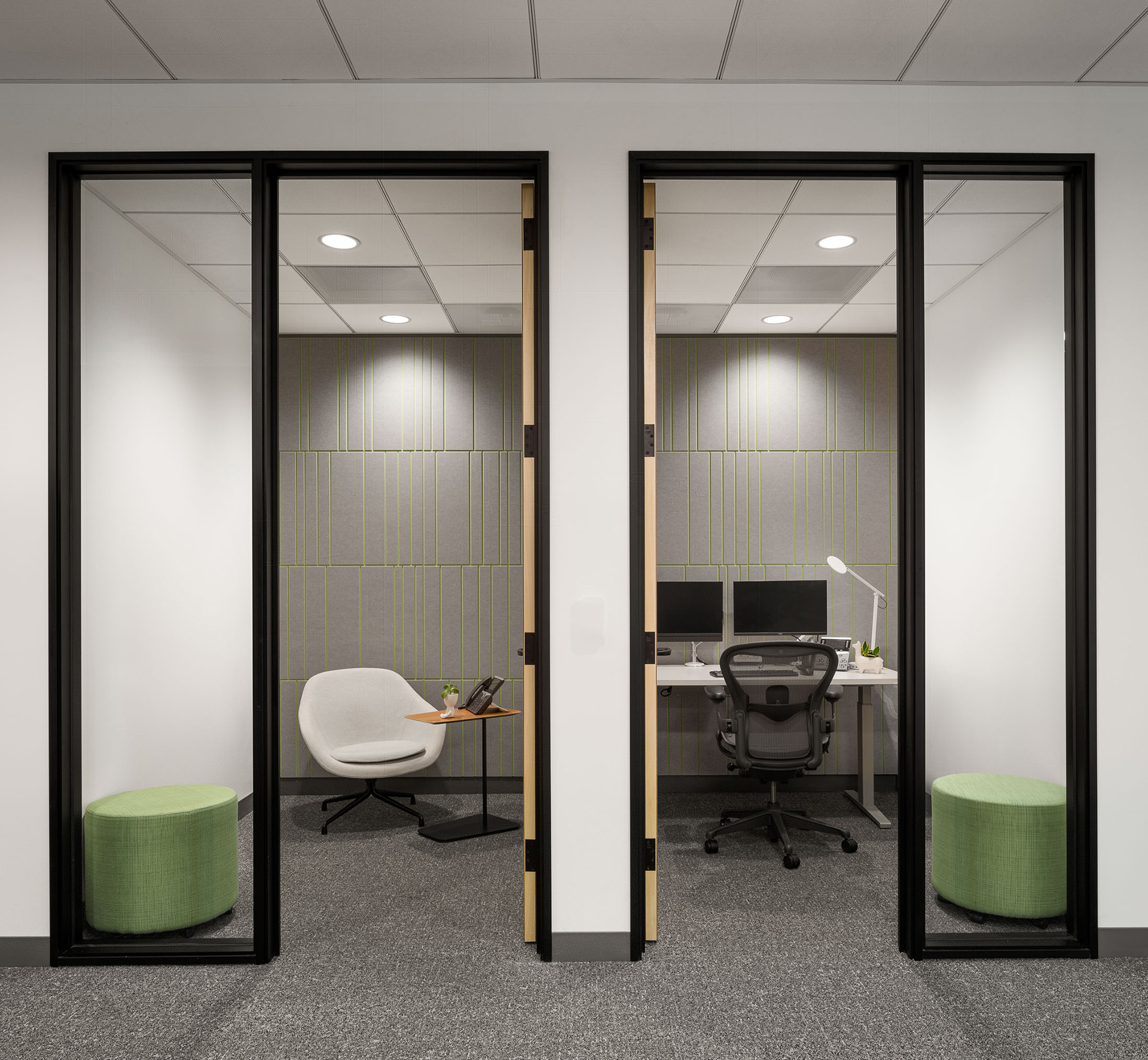 Two small, modern office spaces divided by a glass panel. Each office has a door with a tall window. The left office contains a white chair and green ottoman. The right office is equipped with a desk, task chair, dual monitors, keyboard, mouse, desk lamps. Both offices feature an acoustic felt wall panel for sound absorption.