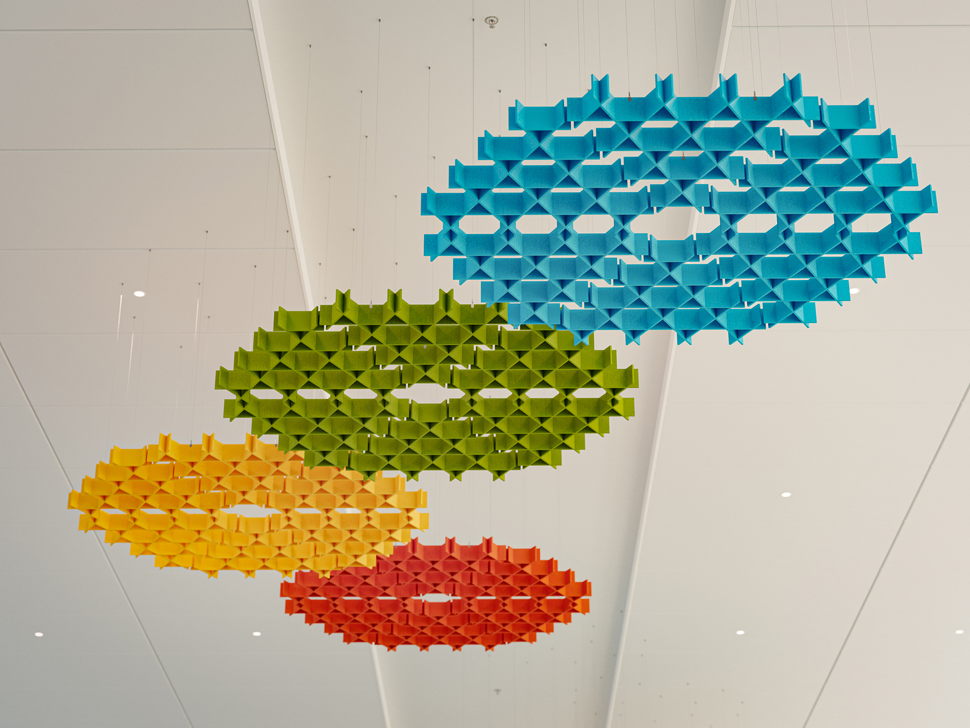 Colorful geometric acoustic felt sculptures—blue, green, yellow, and orange—hang from the ceiling in a white modern space. Each sculpture is a large, circular shape made of interlocking pieces, arranged in a row.