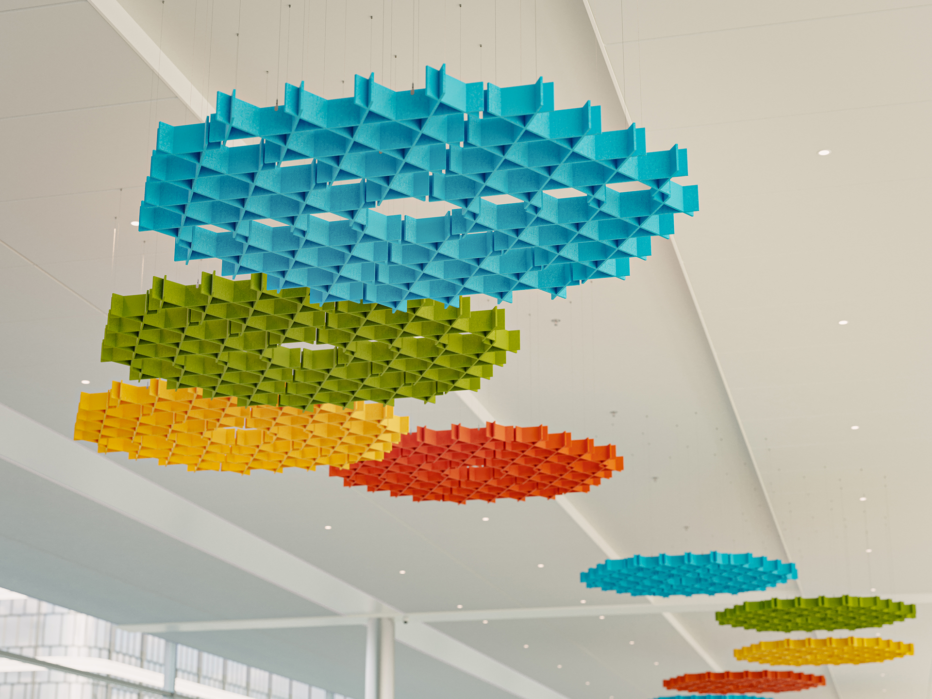 Colorful geometric acoustic felt sculptures, including blue, yellow, green, orange, and red pieces, hang from the ceiling in a modern building with white walls and large windows.