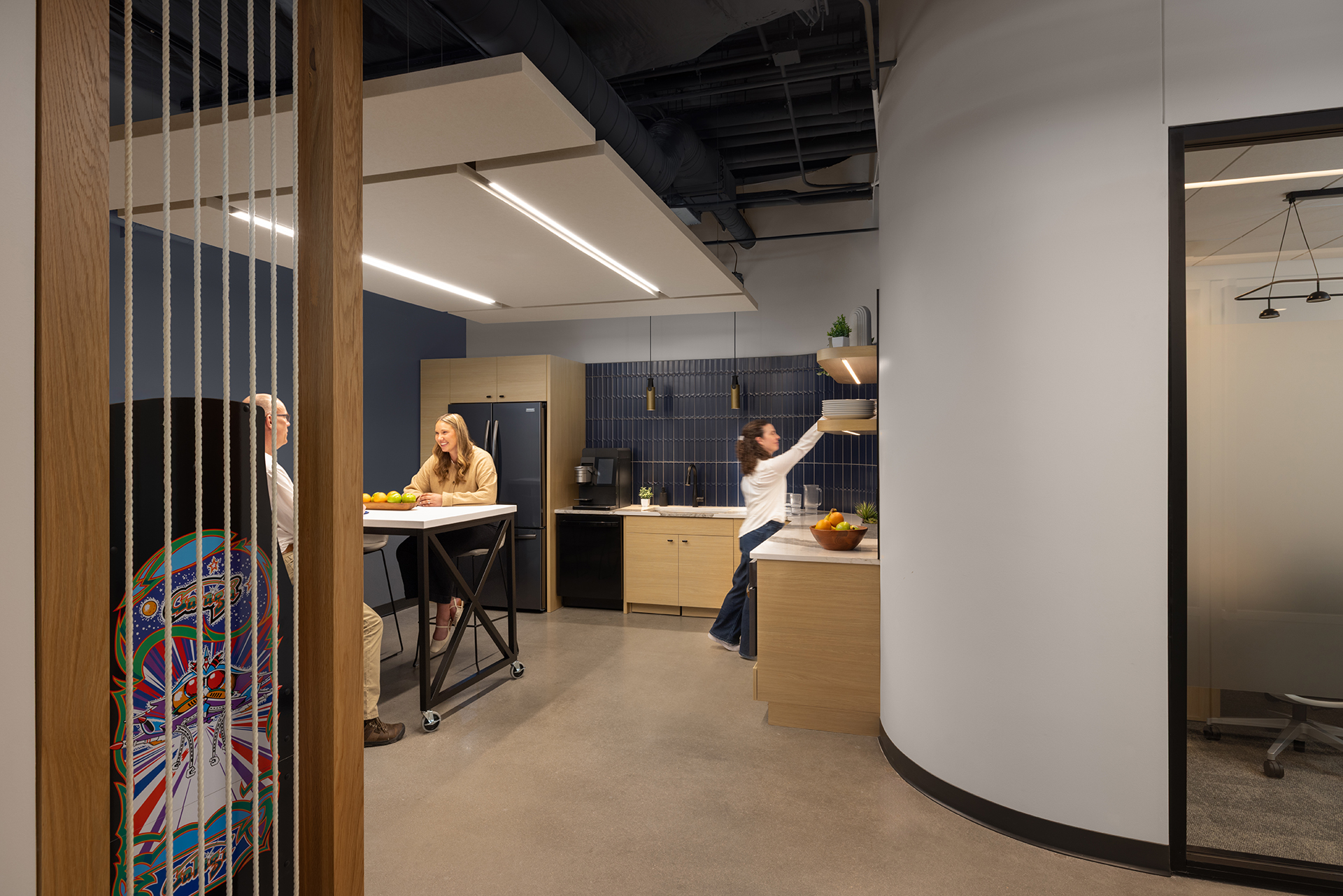 A modern office kitchen with three people: two sitting at a table talking and one standing at a counter. The kitchen has wooden cabinets, a black fridge, fruit bowls, and visible ceiling pipes.
