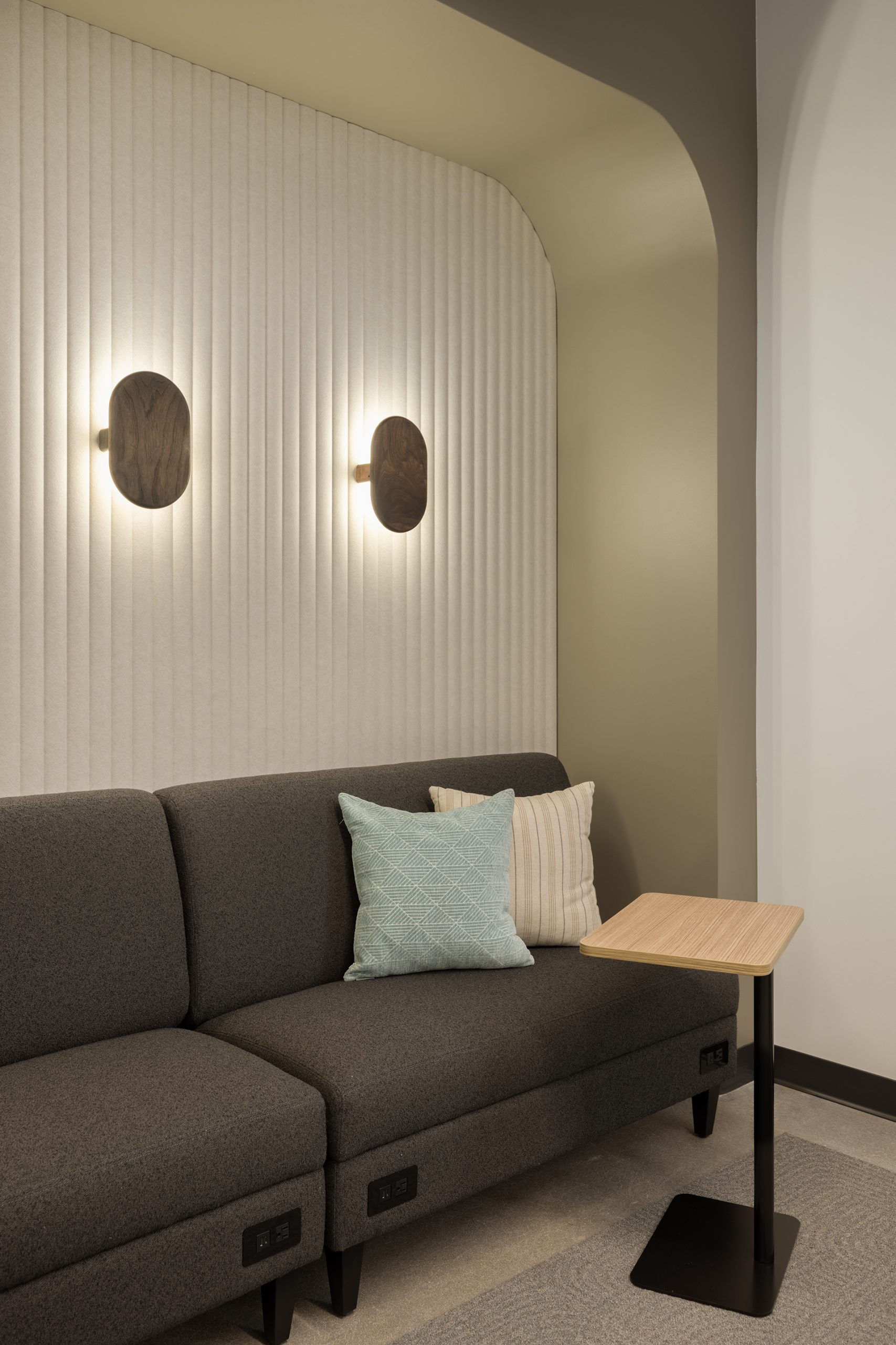 Modern lounge area with a dark gray sofa, two decorative pillows, a small light wood side table, and two oval wall sconces on a textured cream acoustic felt wall. The space features neutral colors and soft lighting.