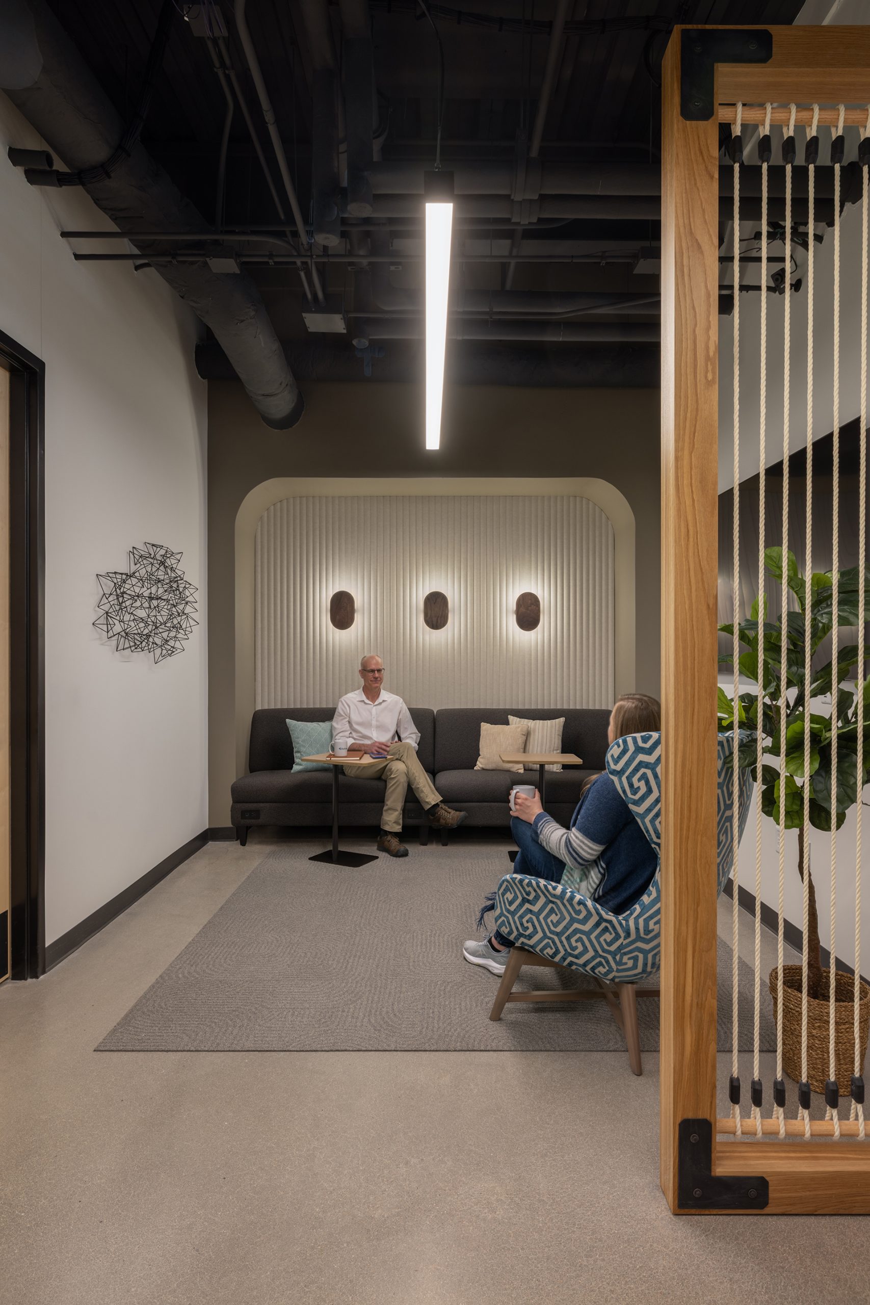 A modern office lounge with two people sitting and talking. The space features a gray sofa, patterned chair, acoustic felt patterned wall, plants, and soft lighting under an exposed ceiling with black ductwork.