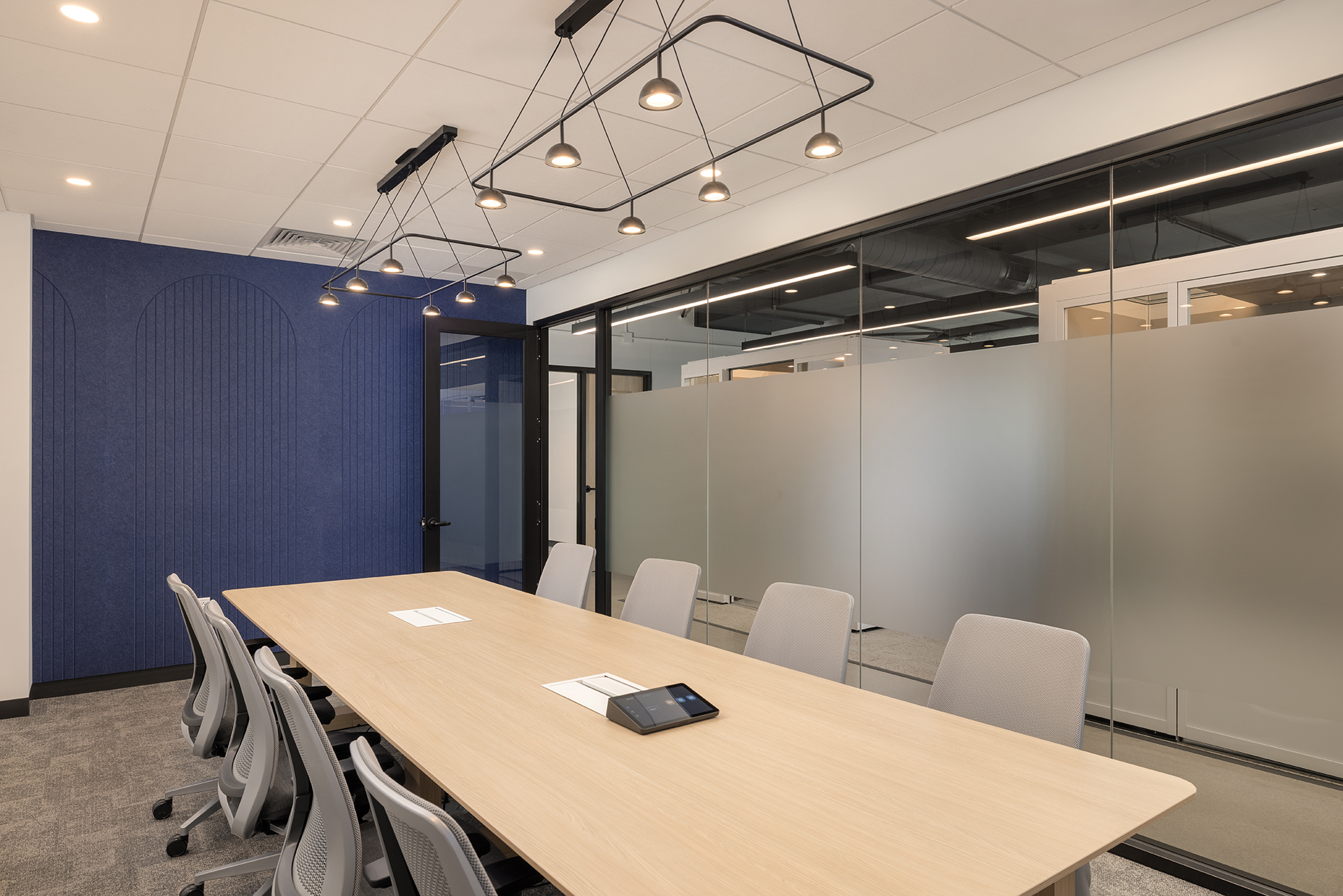 A modern conference room with a long wooden table, ten gray chairs, a blue acoustic felt accent wall, frosted glass partitions, and contemporary pendant lighting. A speakerphone and papers are on the table.