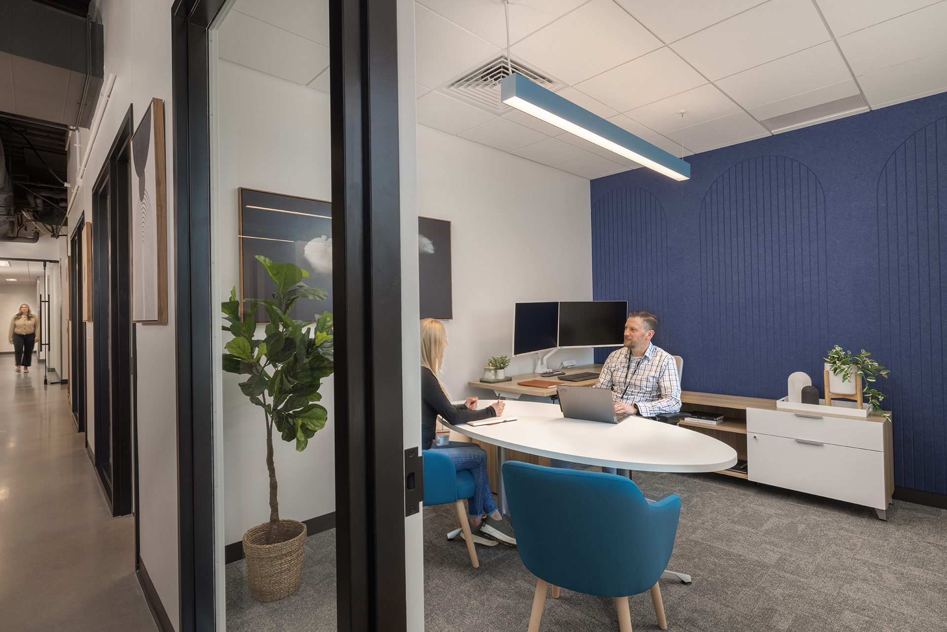 A man and a woman sit across from each other at a desk in a modern office. The room has blue chairs, a large plant, two monitors, and a blue acoustic felt accent wall. A person walks down the hallway outside the office.