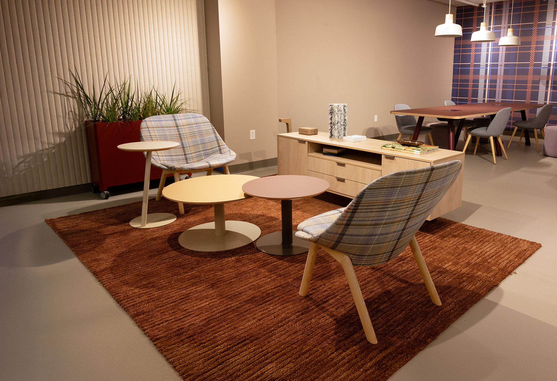 A modern lounge area with two plaid chairs, three round tables, a brown rug, a wooden console, an acoustic felt accent wall, and a red planter with tall green plants. In the background, a dining table with gray chairs and pendant lights is visible.
