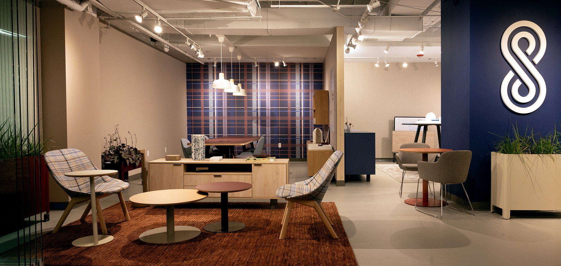 A modern office lounge with plaid chairs, round tables, a brown rug, pendant lights, potted plants, and a blue accent wall with a white abstract logo. The area has neutral tones and a plaid-patterned wall in the background.