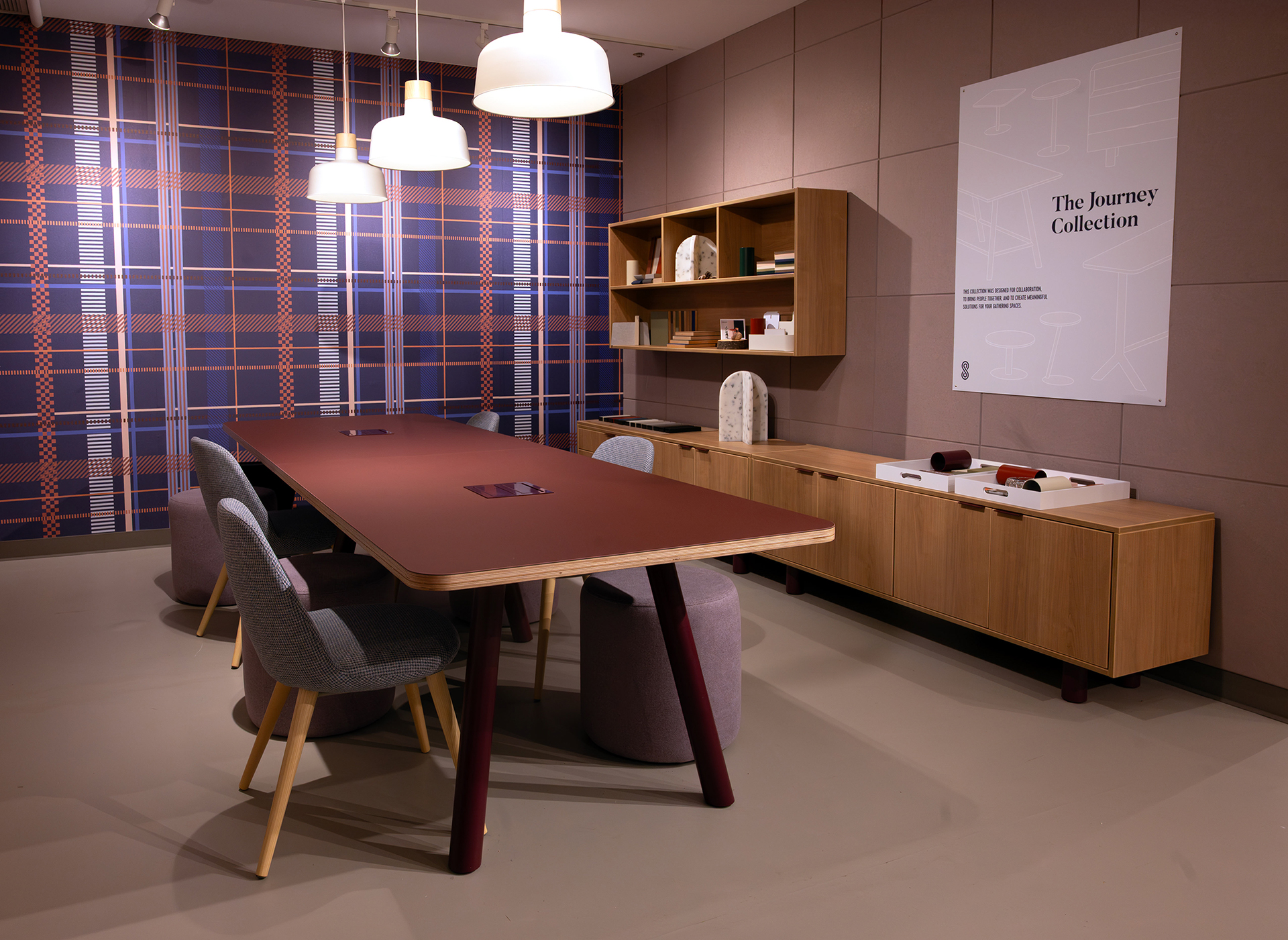 A modern meeting room with a long table, gray and mauve chairs, a wooden cabinet, shelves with decor, and pendant lights. The wall features a blue plaid pattern and a large poster reading "The Journey Collection" set against an acoustic felt accent wall.