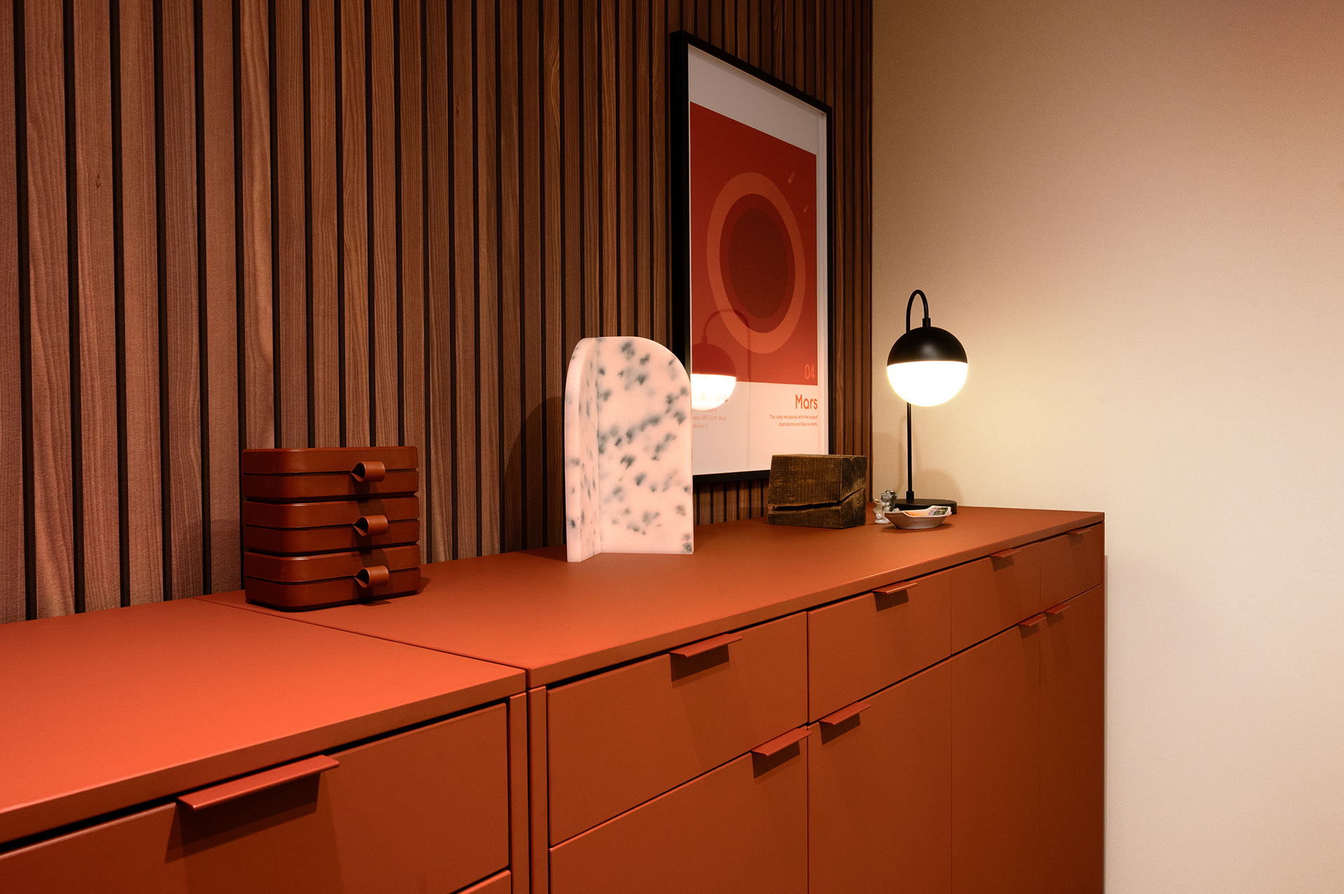 A modern interior featuring matte orange cabinets, a ribbed acoustic felt woodgrain wall, a framed Mars poster, a marble decor piece, stacked boxes, a small lamp, and a tray with items, all warmly lit.