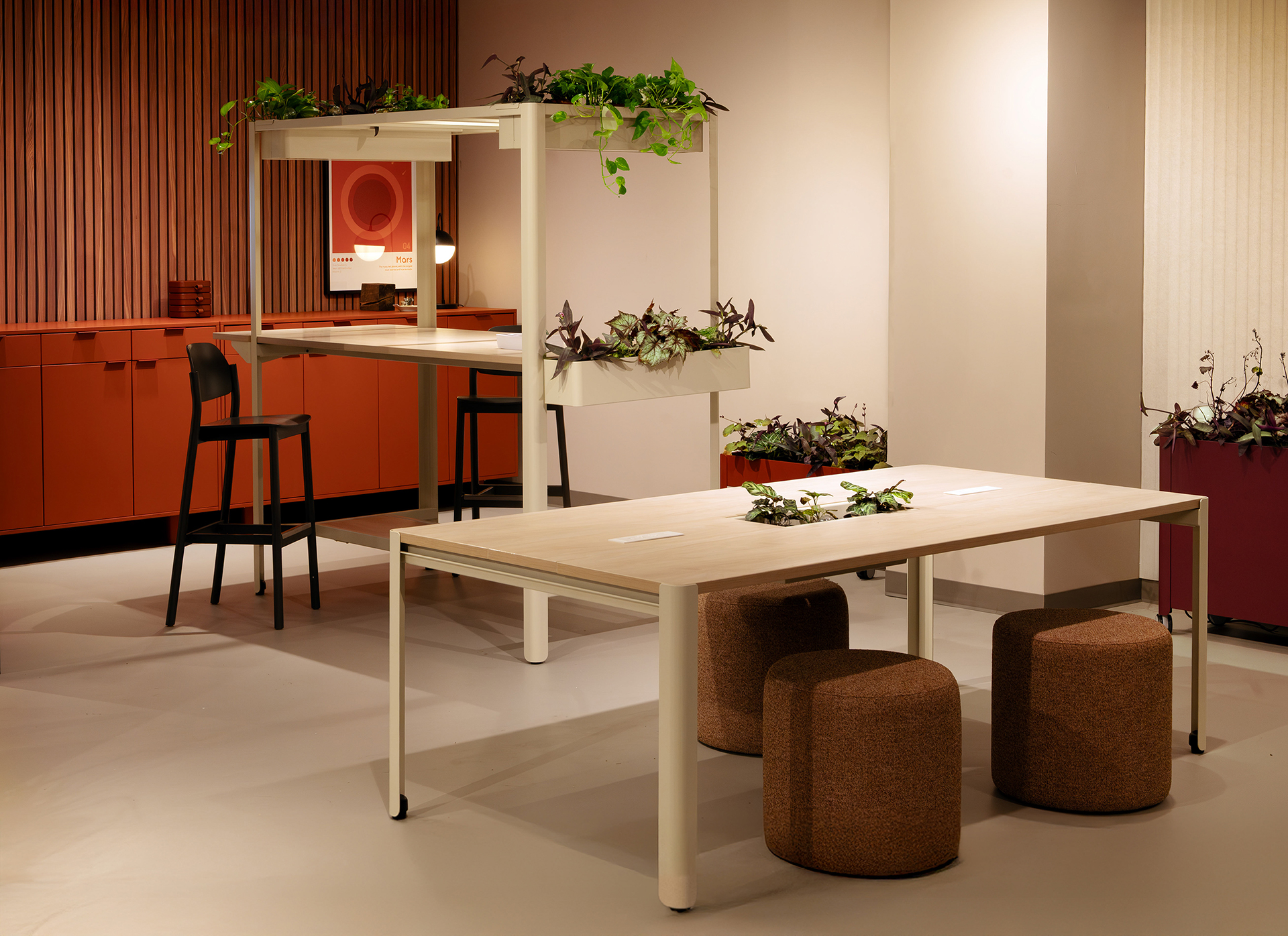 Modern workspace with light wood tables, brown pouf seats, and a high chair. Green plants decorate the tables and a planter in the background. Red cabinets, a woodgrain acoustic felt accent wall, and warm lighting create a cozy, inviting atmosphere.