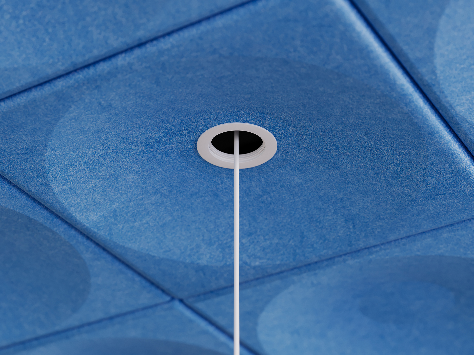 A blue acoustic felt ceiling tile with a hole cut into it where lighting hardware fits and and a light fixture hangs down below the soft architecture installation.