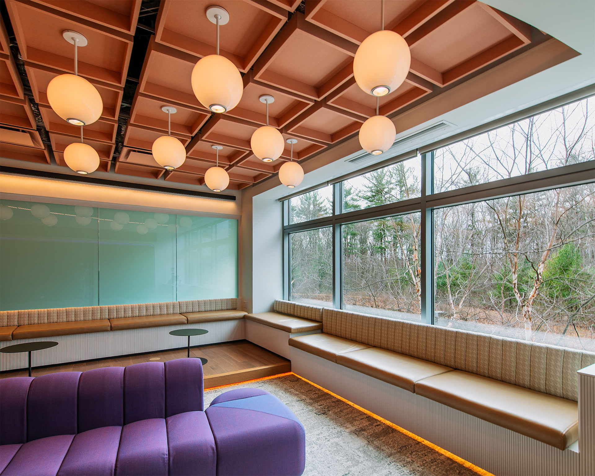 Modern lounge area with a purple sofa, long window seating, round ceiling lights, acoustic felt ceiling clouds, and large windows overlooking trees and a natural outdoor landscape.