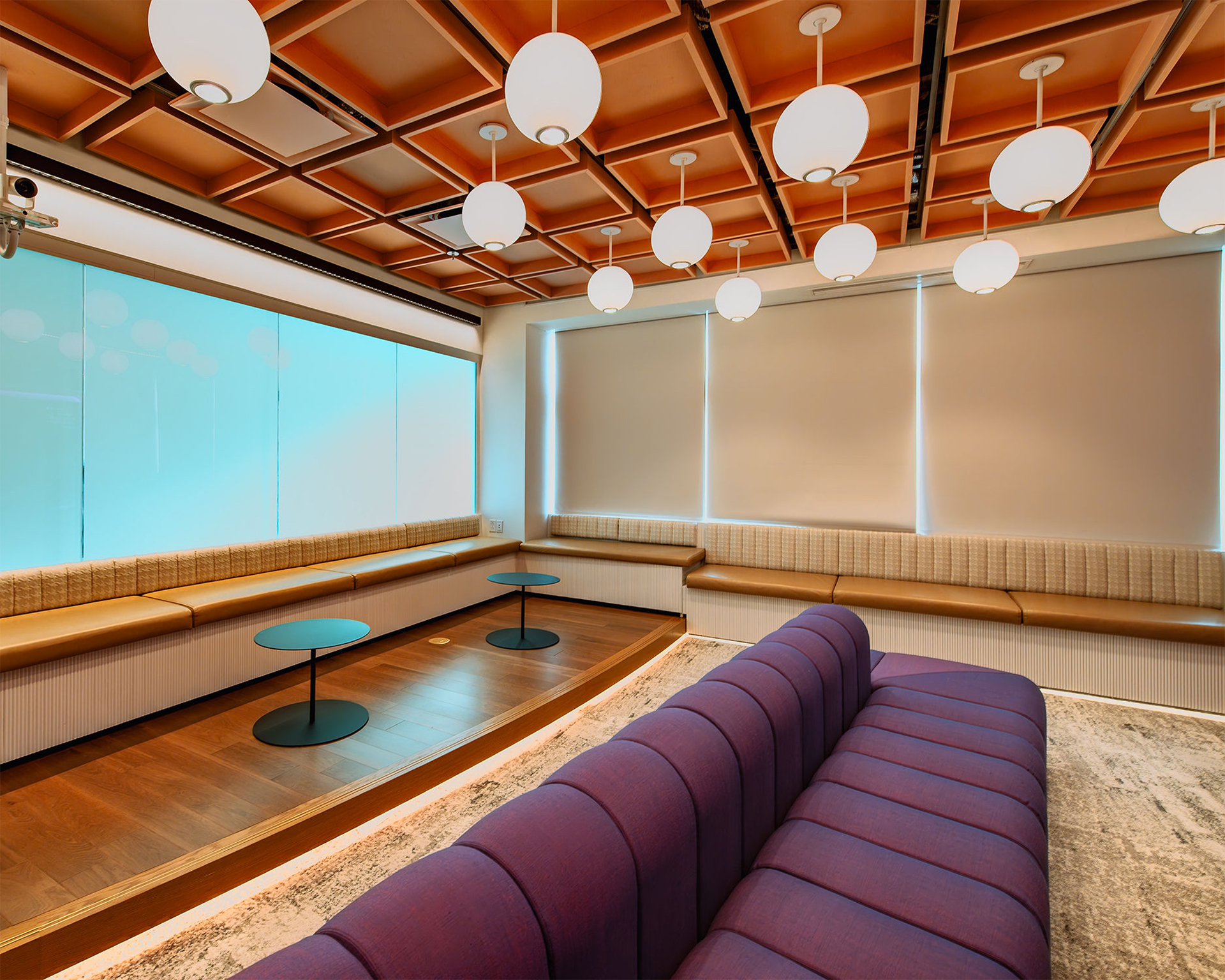 A modern lounge features a purple sofa, round blue tables, built-in bench seating, large windows with blinds, and an acoustic felt ceiling with spherical pendant lights.