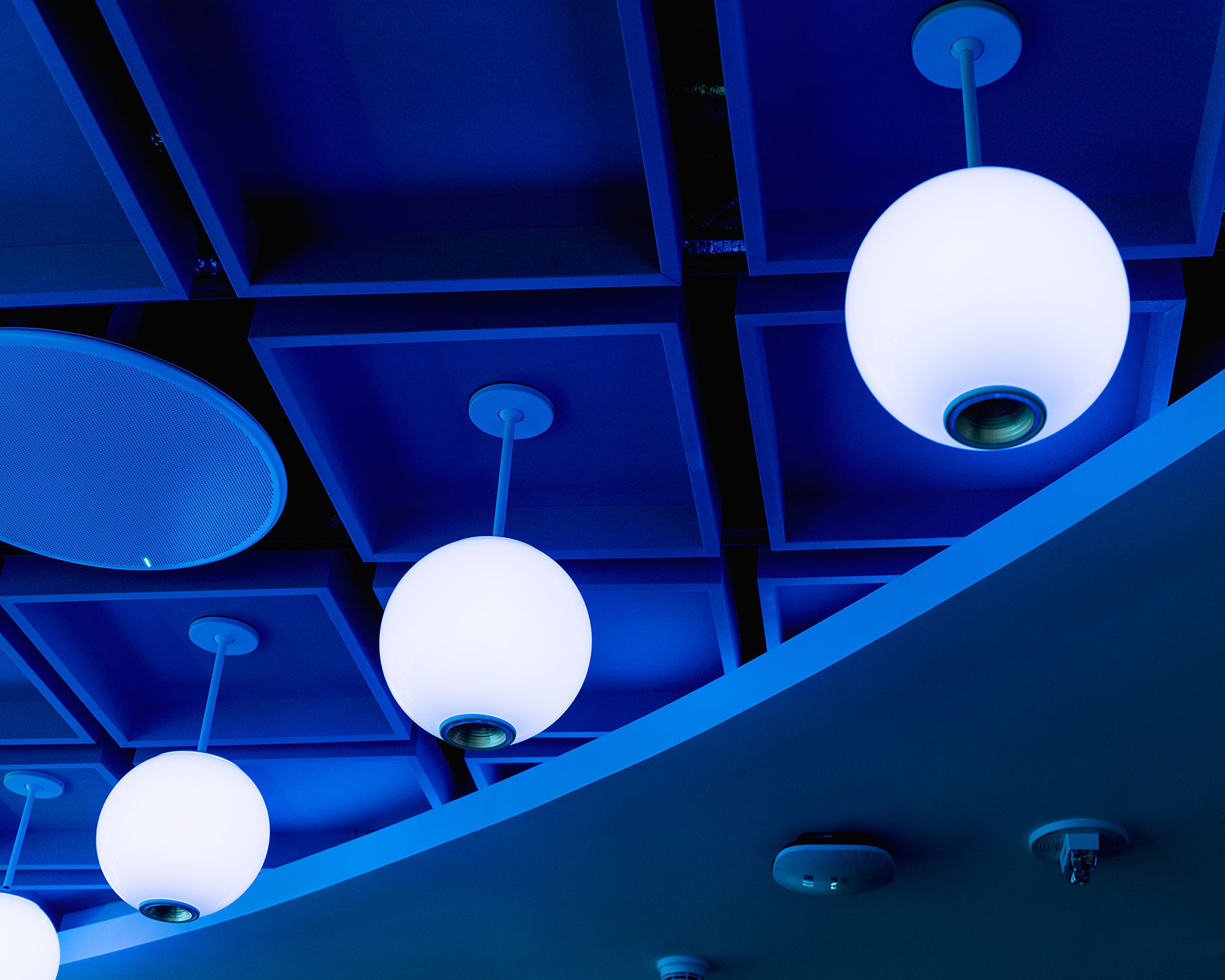 A row of round, glowing pendant lights hangs from a grid-patterned acoustic felt ceiling with geometric shapes and curved architectural lines.
