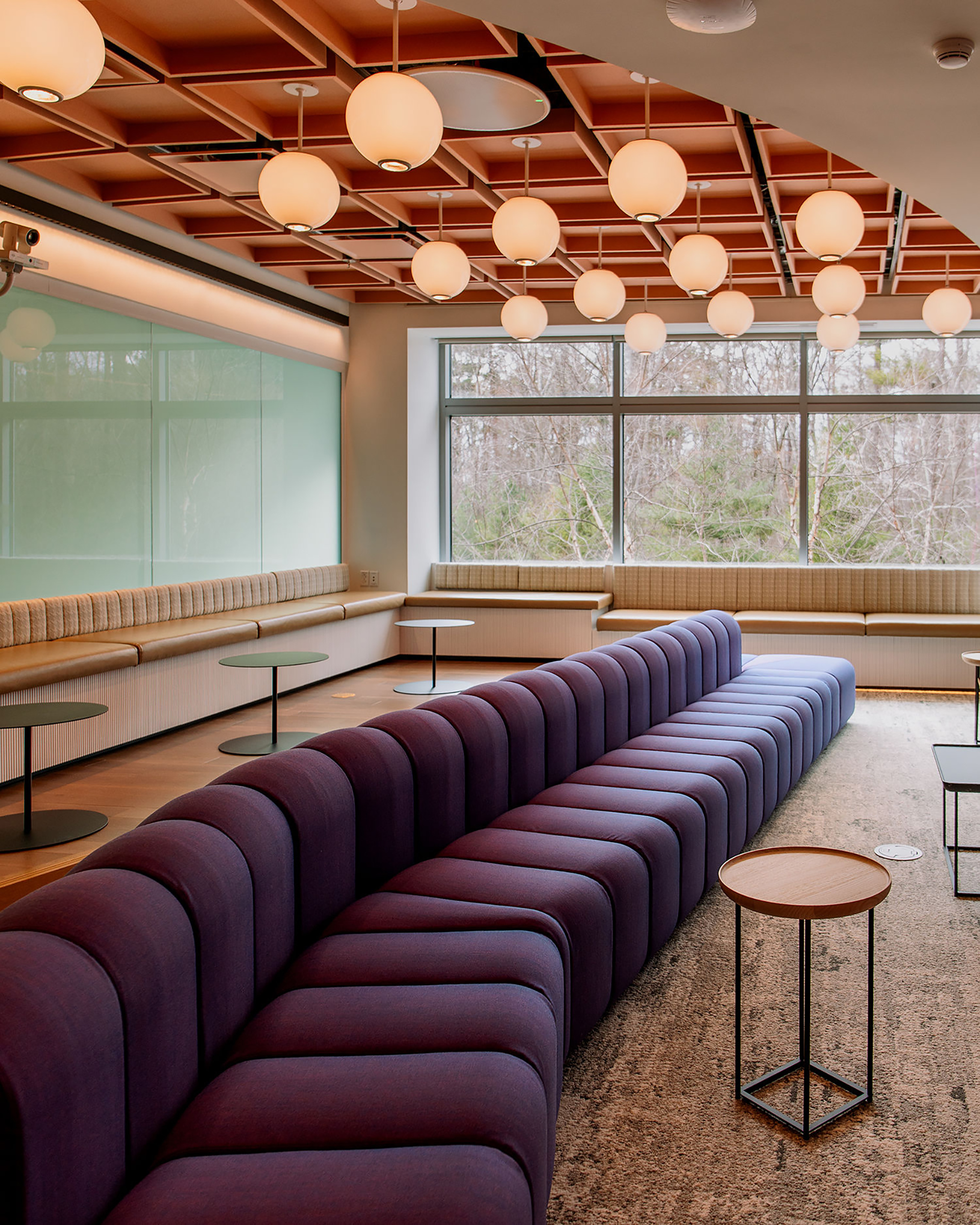 Modern lounge with a long, curved purple sofa, round wooden tables, globe pendant lights on an acoustic felt cloud ceiling, built-in beige bench seating, large windows, and a view of trees outside.