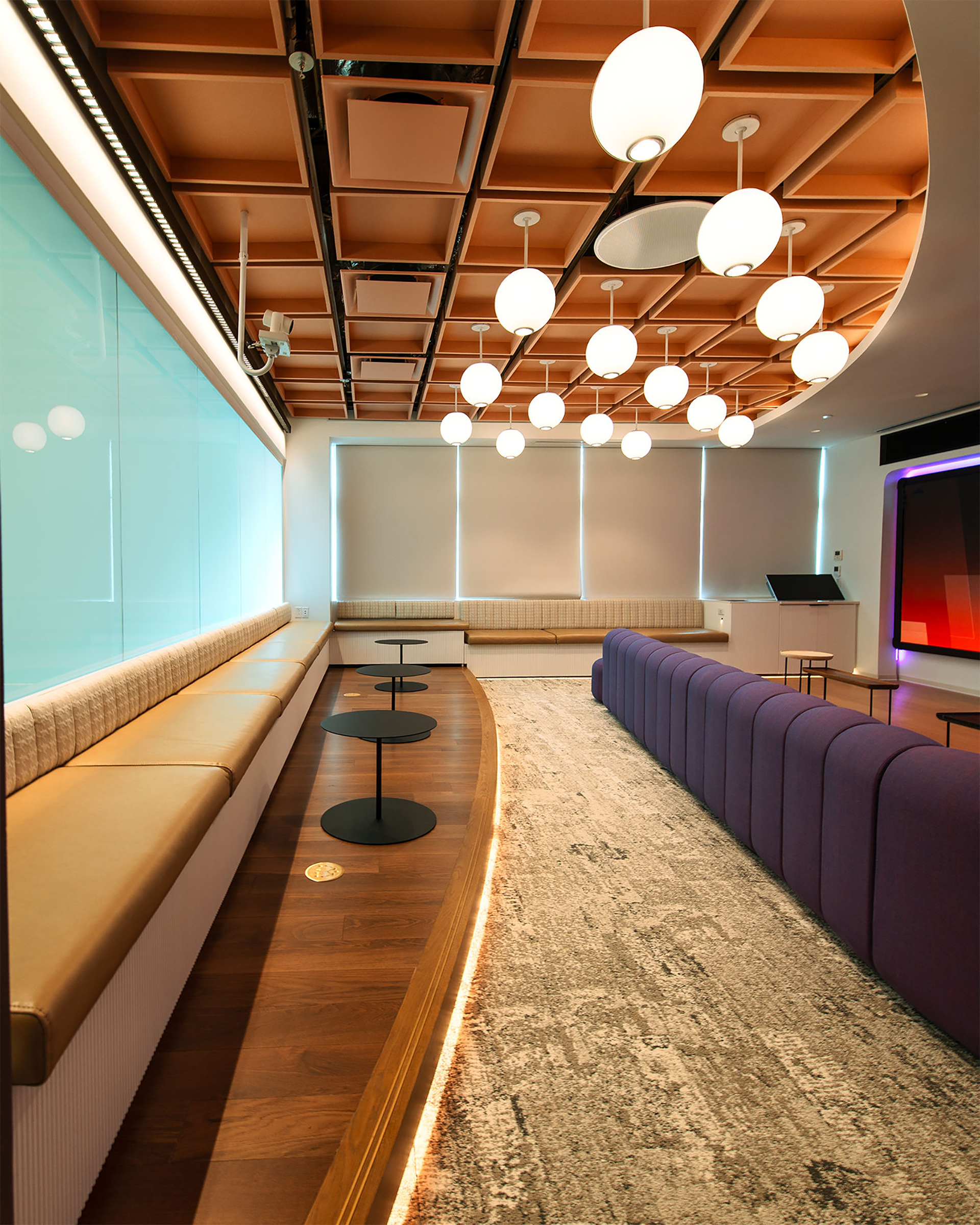 A modern lounge with tan bench seating, round black tables, a purple sofa, a textured rug, and a ceiling with globe pendant lights and square acoustic felt ceiling clouds. Large windows line the back wall, and lighting adds a warm ambiance.