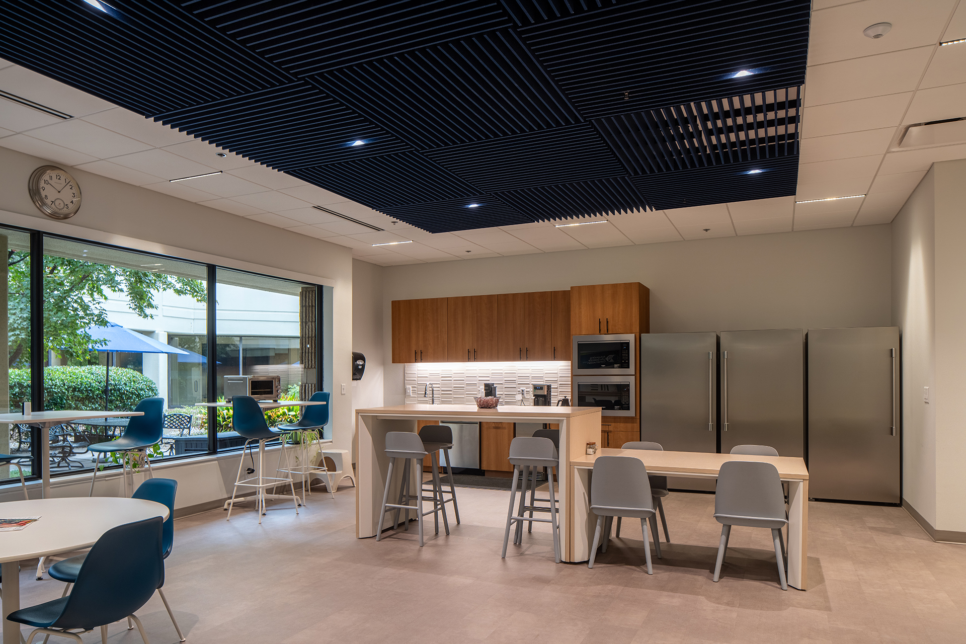 Modern office kitchen with wooden cabinets, stainless steel appliances, tables, and gray chairs. Large windows provide a view of an outdoor patio with trees and tables. The ceiling features dark blue acoustic felt frames.