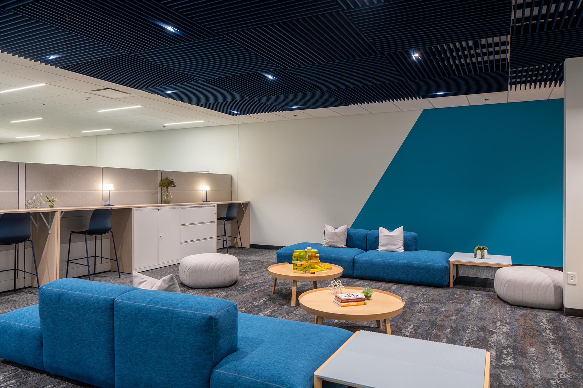 A modern office lounge with blue couches, light wood tables, white poufs, and a blue accent wall. In the background are high chairs and workstations under a ceiling with dark slatted panels and built-in lights.