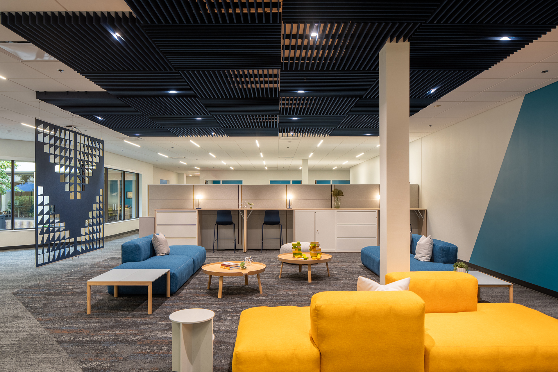 Modern office lounge with yellow and blue sofas, round coffee tables, and a geometric dark blue acoustic felt ceiling design. An acoustic felt screen with triangular cutouts separates the space, and high desks with stools are in the background.