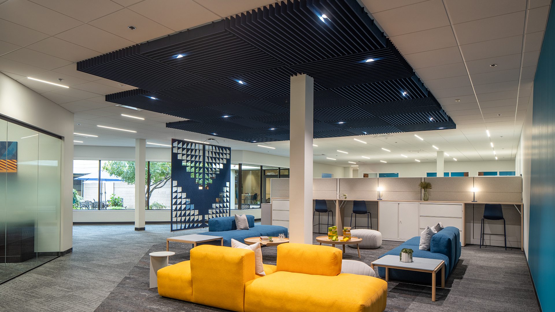 Modern office lounge with bright yellow and blue sofas, accent tables, and a decorative acoustic felt ceiling frame. Large windows provide natural light, and glass walls separate different areas. An acoustic felt screen provides privacy and indoor plants add a touch of greenery.