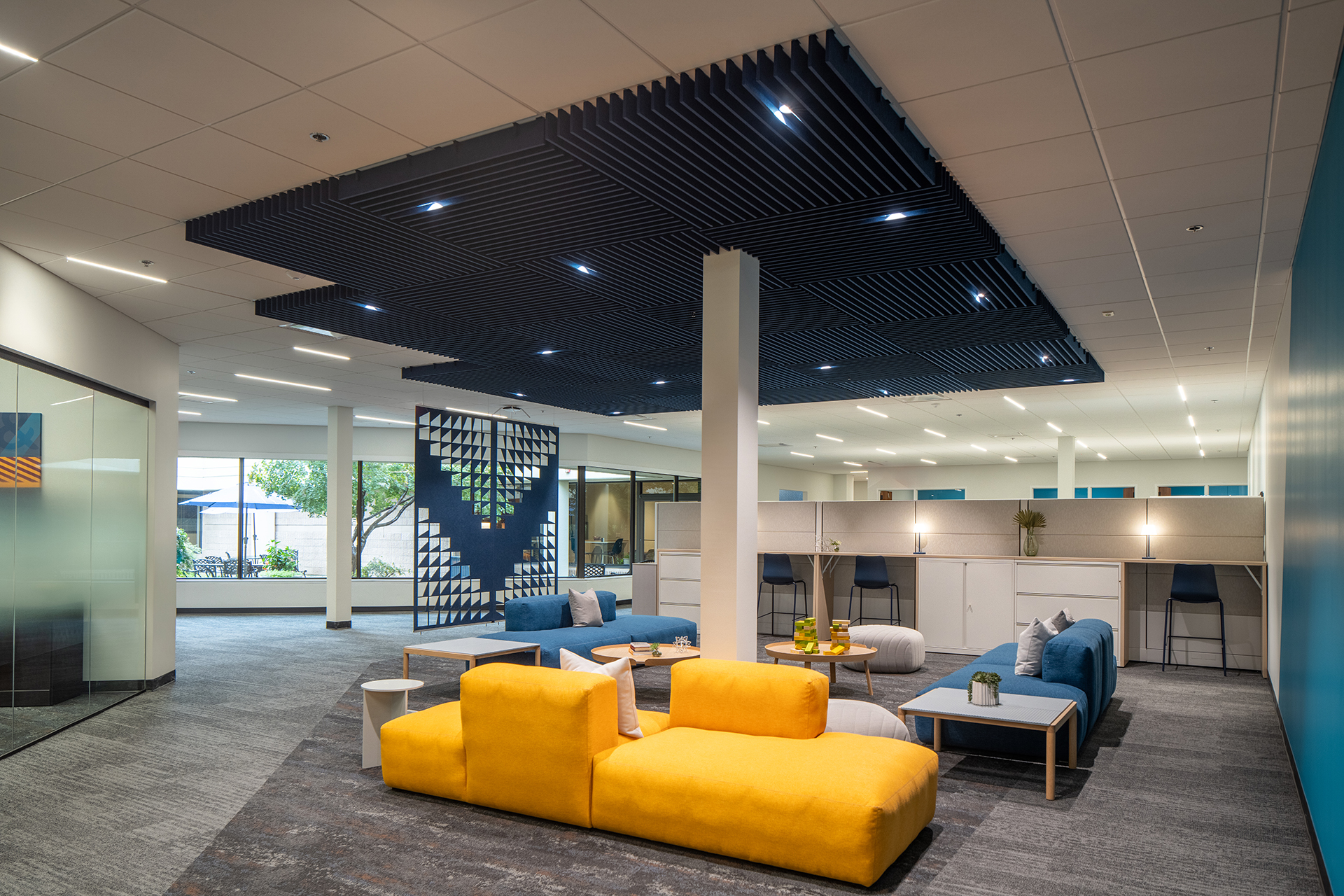 Modern office lounge with bright yellow and blue sofas, accent tables, and a decorative acoustic felt ceiling frame. Large windows provide natural light, and glass walls separate different areas. An acoustic felt screen provides privacy and indoor plants add a touch of greenery.