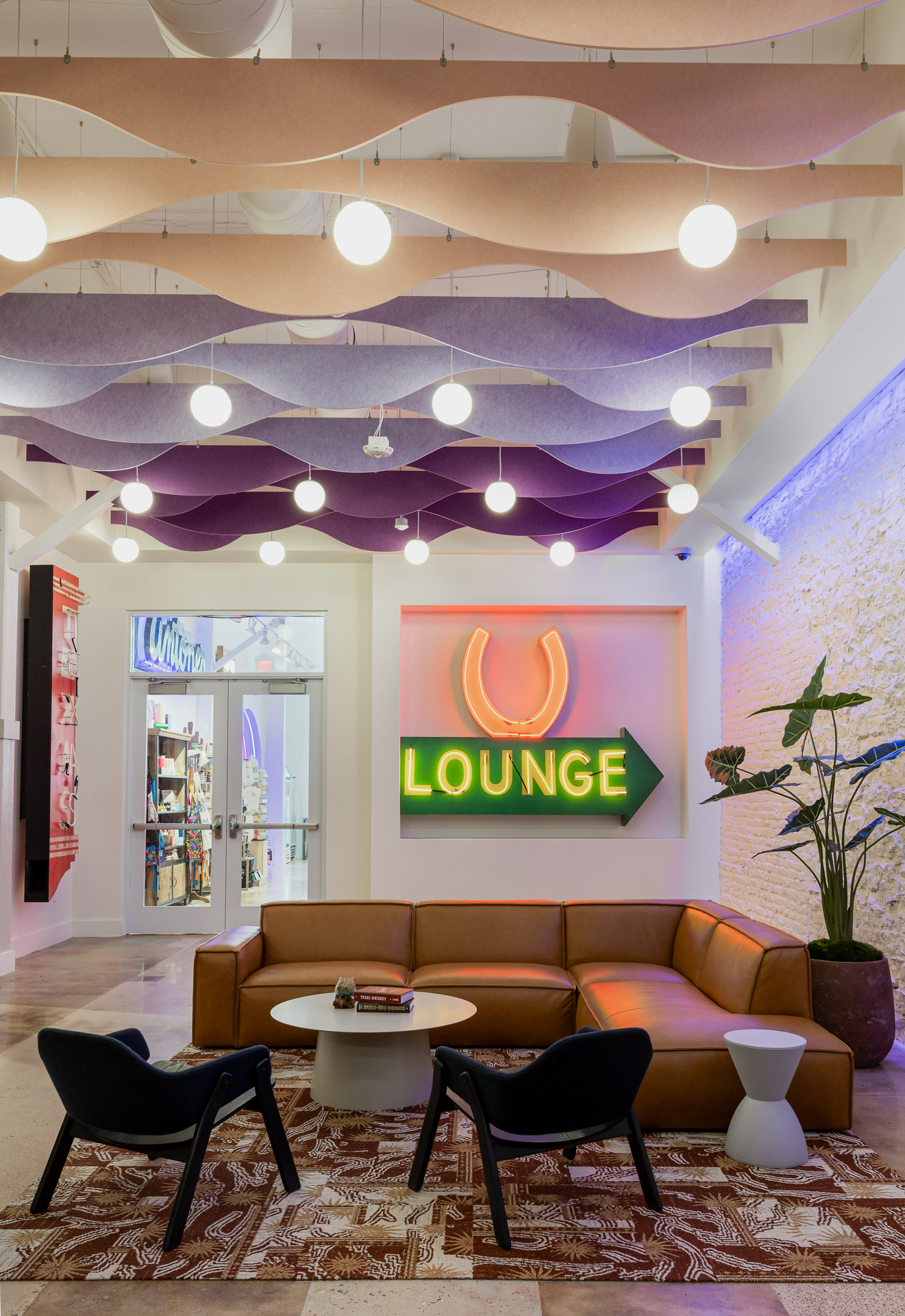 Contemporary lounge with a brown leather sectional sofa, two black chairs, a white coffee table, and a neon "LOUNGE" sign on the wall. Wavy acoustic felt baffles and round lights hang from the ceiling. A patterned rug and plant add decor.
