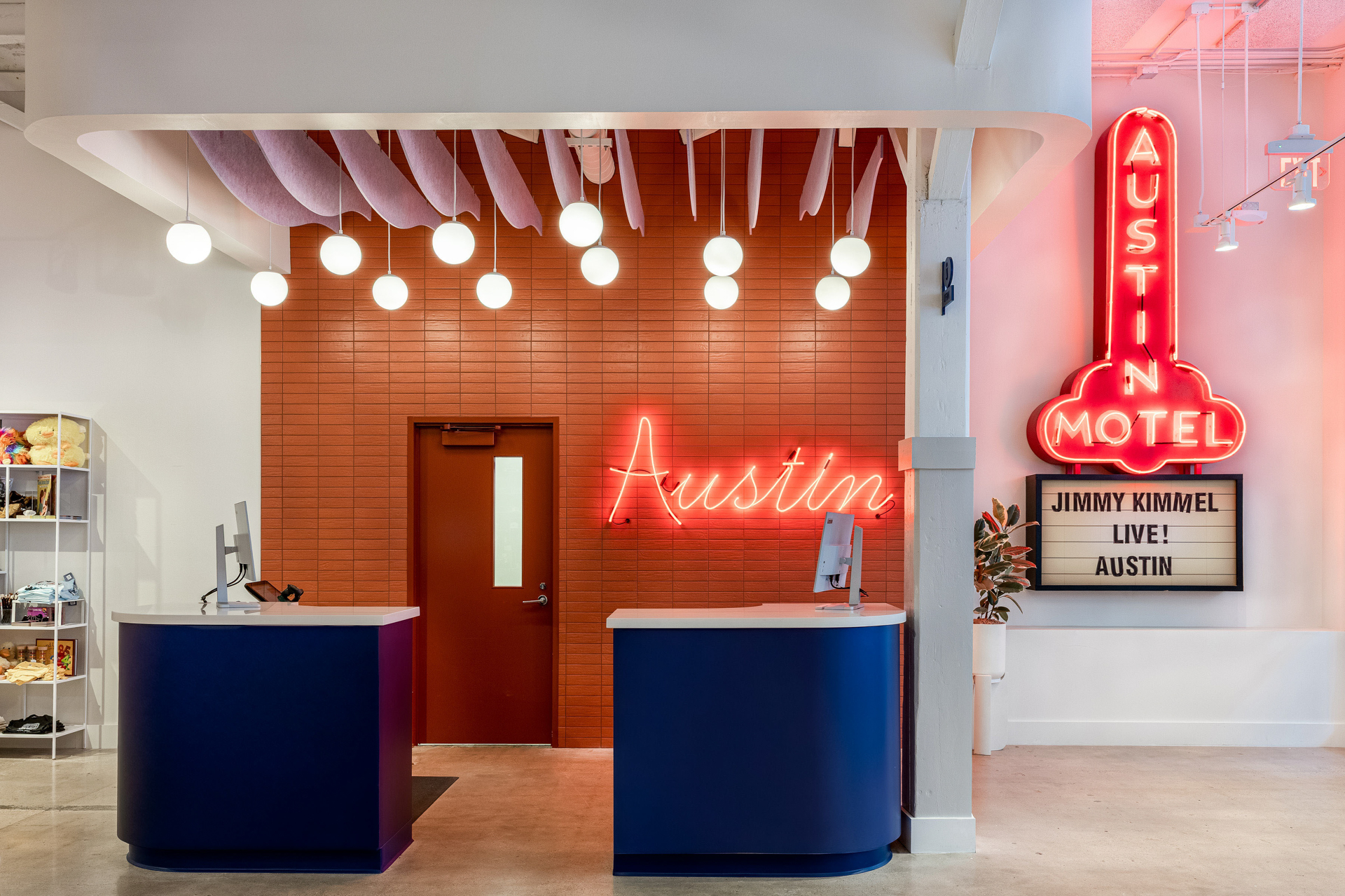 Hotel lobby with blue desks, a red tiled wall, neon "Austin" sign, and a large pink neon "Austin Motel" sign. Spherical lights hang from the ceiling which has acoustic felt baffles; a shelf with items is visible on the left.