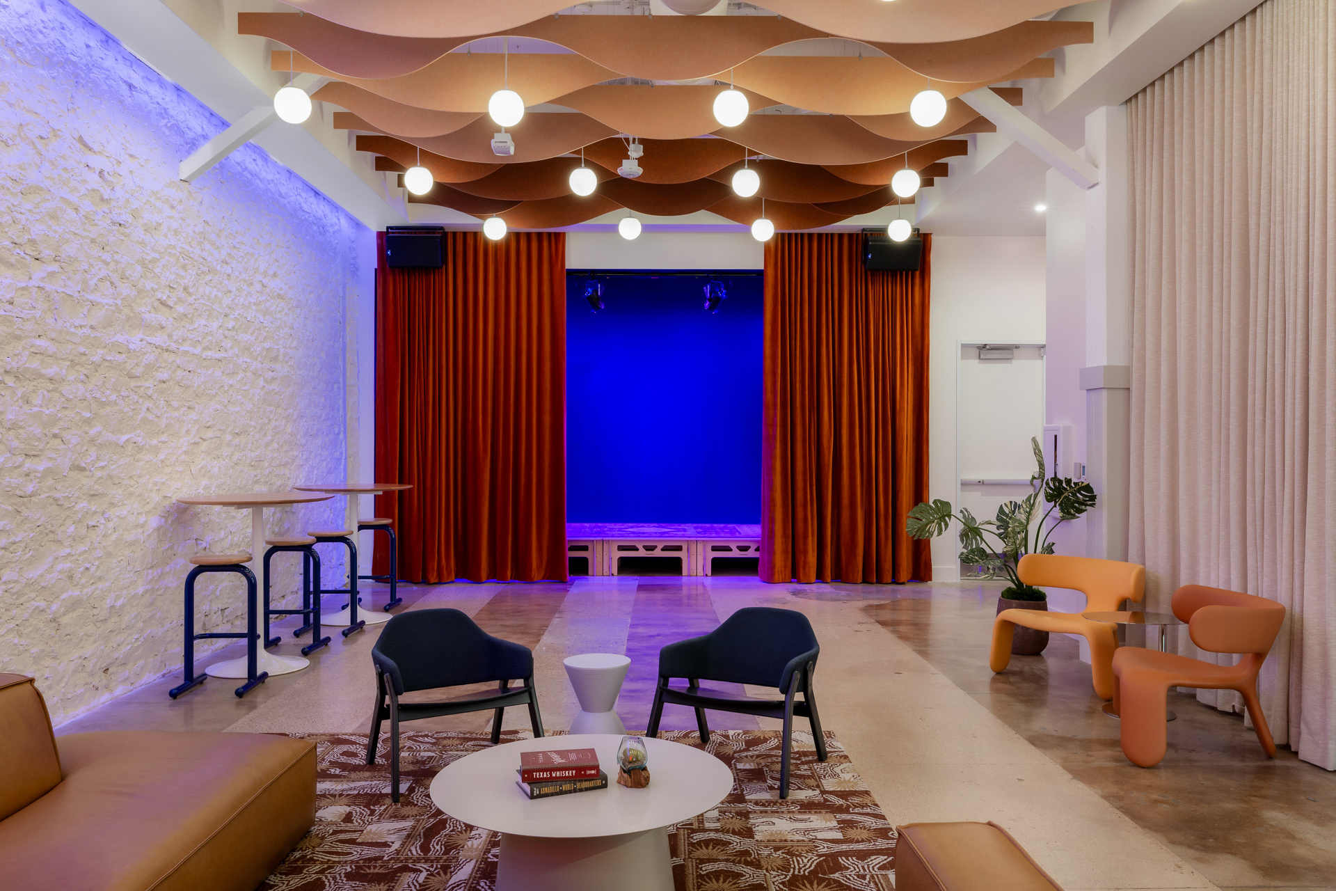 A modern lounge with a brown sofa, black chairs around a white coffee table, a stage with red curtains and blue lighting, wavy acoustic felt ceiling baffles, plants, and a textured white wall.