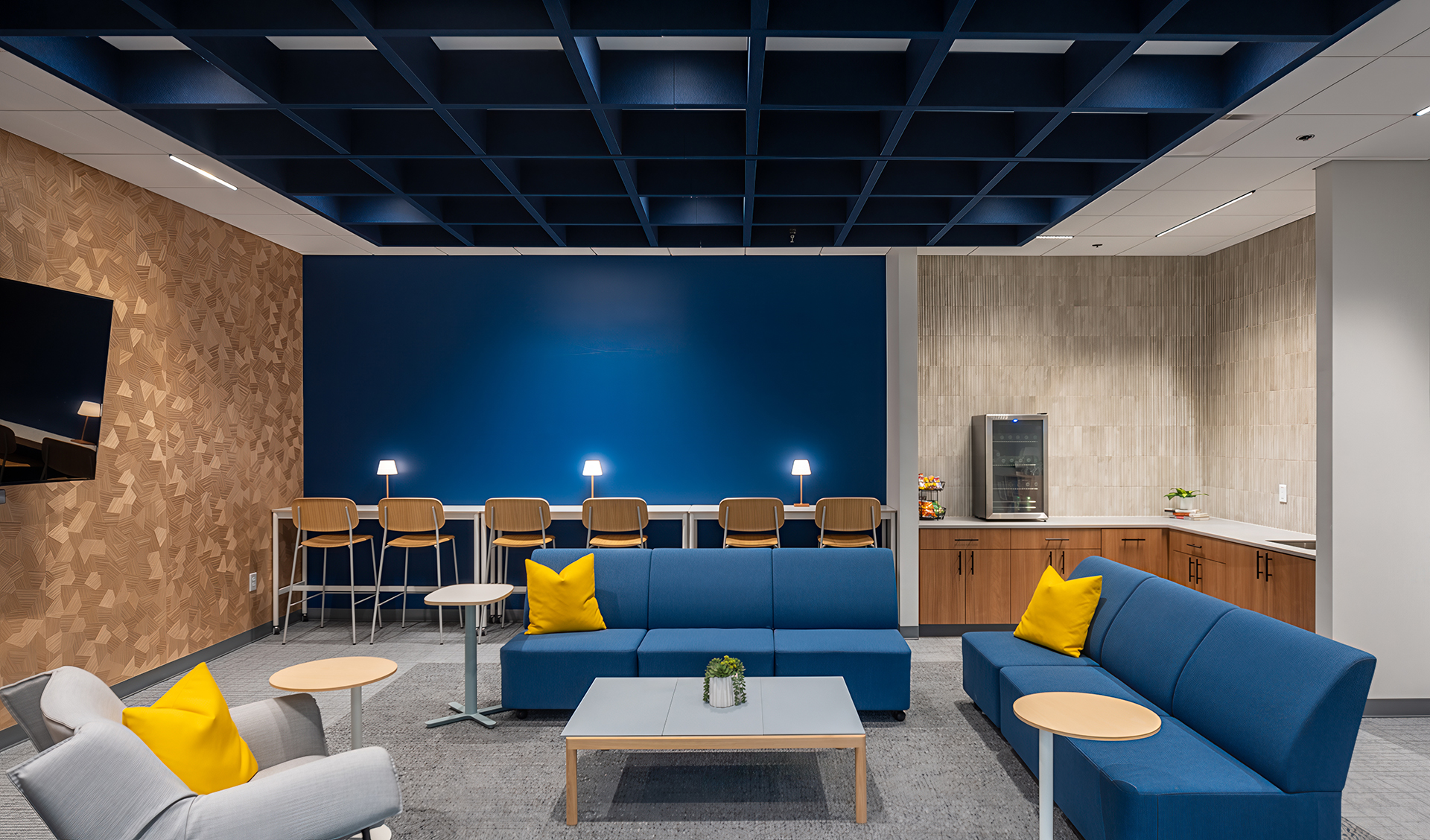 Modern lounge with blue sofas, yellow pillows, armchairs, a coffee table, a high counter with chairs and lamps, a wood accent wall, and a blue acoustic felt ceiling grid. A water dispenser and cabinets are in the corner.