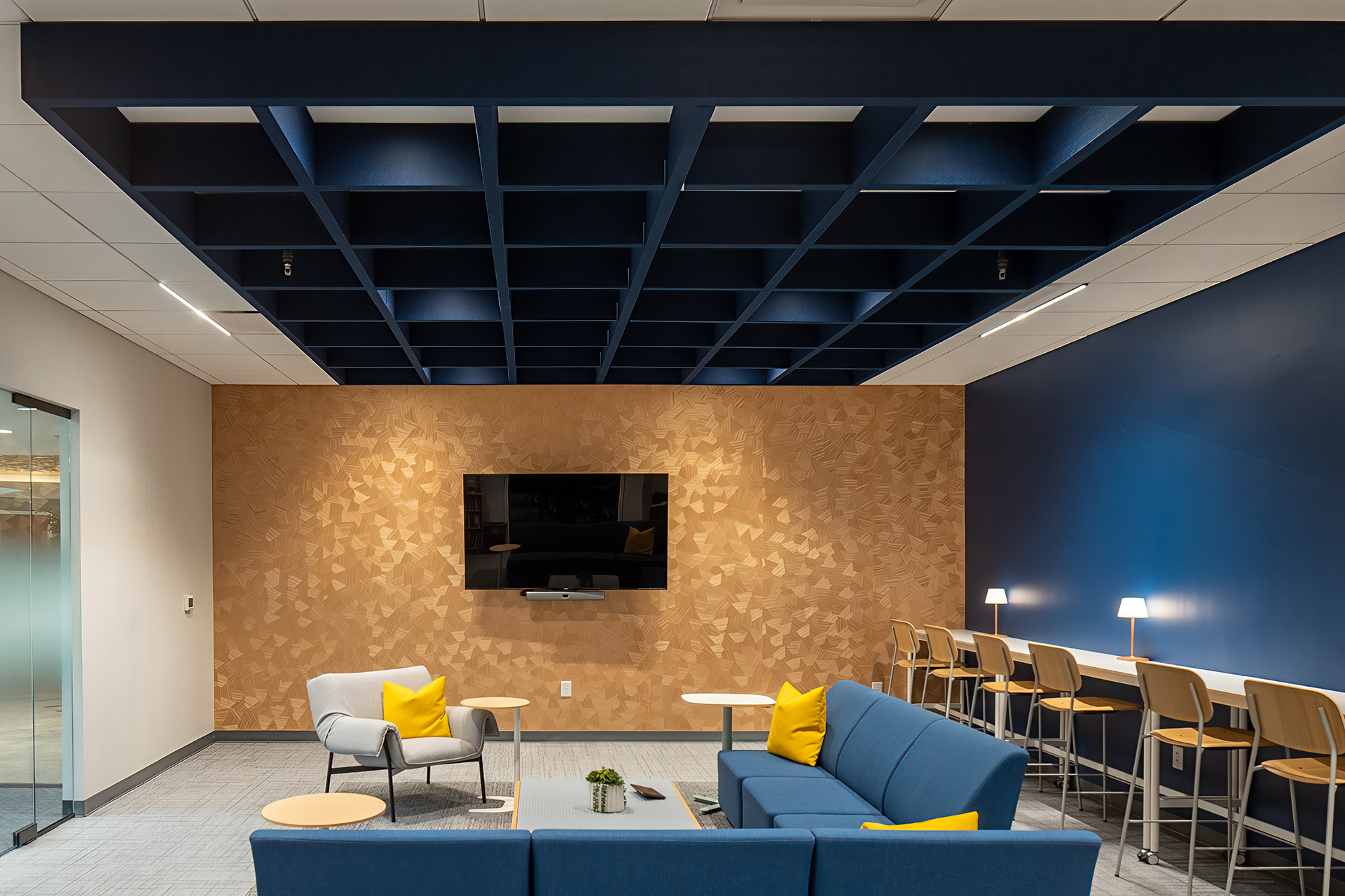 Modern office lounge with blue couches, yellow accent pillows, a wall-mounted TV, a wooden accent wall, high counter with stools and small lamps, and an acoustic felt ceiling with a blue grid design. Glass door visible on the left.