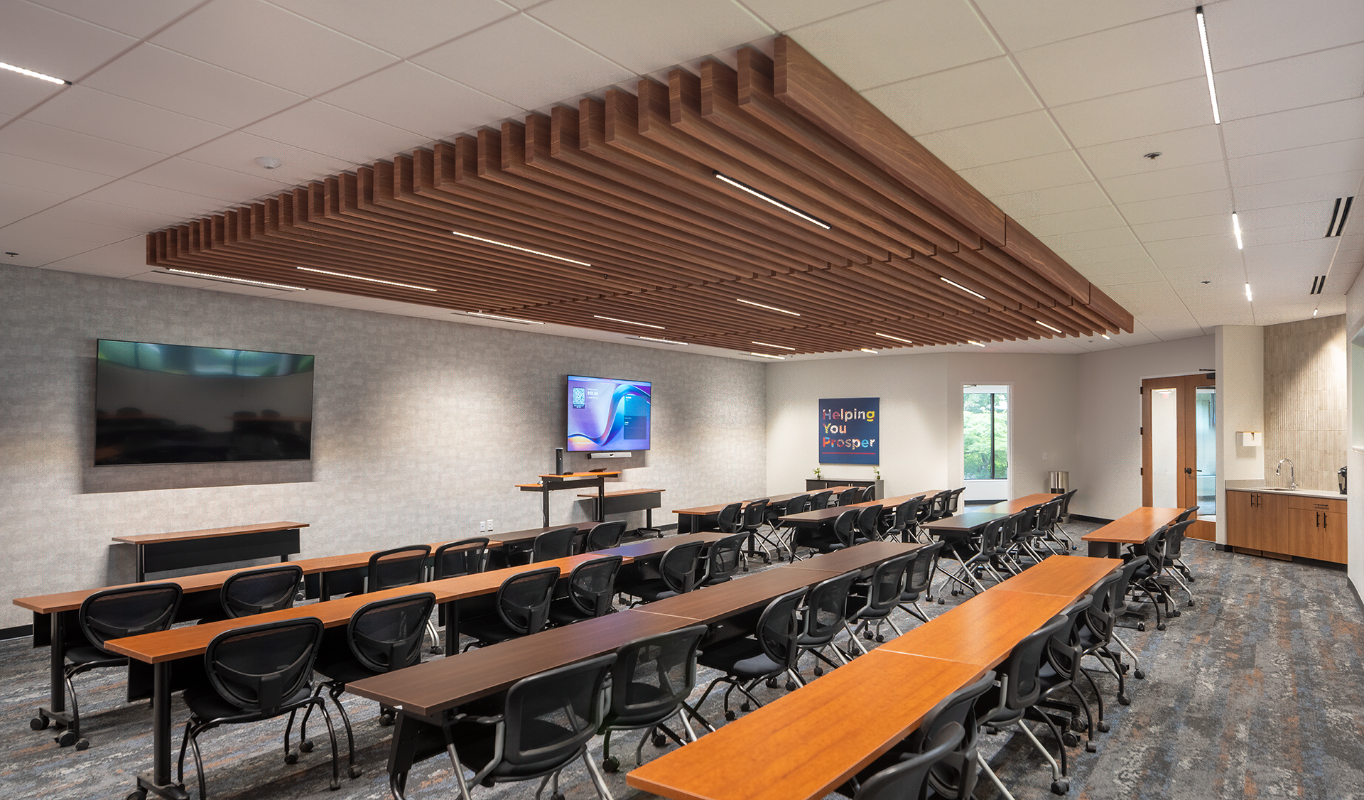 A modern classroom with rows of tables and chairs facing two large wall-mounted screens, a podium, and a sign that reads “Helping You Succeed” on the back wall. The ceiling features acoustic felt woodgrain ceiling baffles.