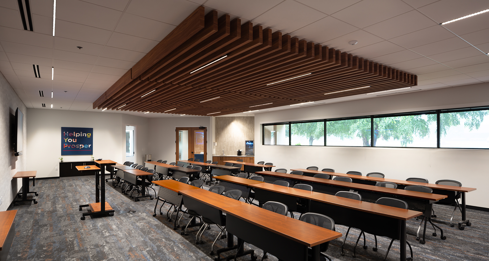 A modern classroom with rows of wooden desks and black chairs, large windows along one wall, a podium at the front, and a colorful "Helping You Prosper" sign on the left wall. The ceiling features acoustic felt woodgrain ceiling baffles.