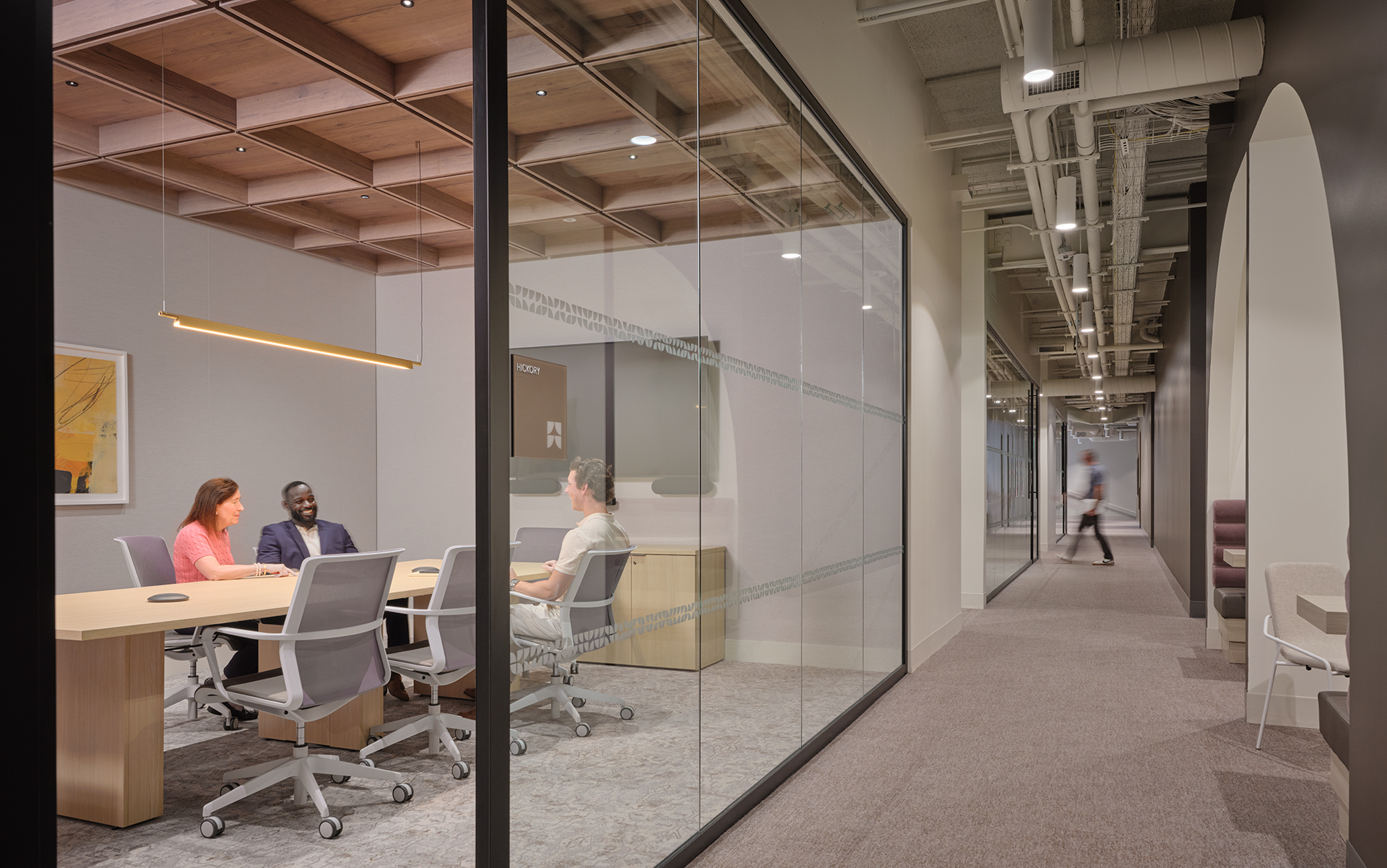 Three people sit around a table in a modern glass-walled conference room. A hallway runs alongside the room, with a blurred figure walking in the distance. The office space looks contemporary and well-lit.