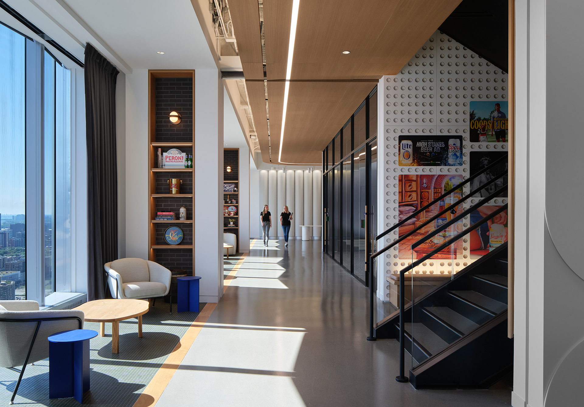 Modern office lounge with large windows, cozy seating, bookshelves, and board games. Two people walk down a bright hallway past a wall decorated with colorful posters and an open staircase. Cityscape visible outside.