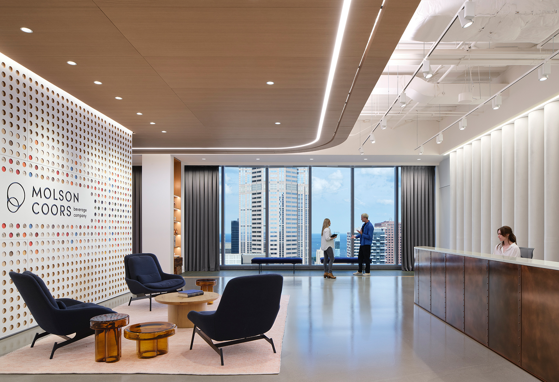 Modern office lobby with a Molson Coors logo on the left wall, stylish seating area, large windows showing city buildings, two people conversing by the window, and a receptionist at a front desk on the right.