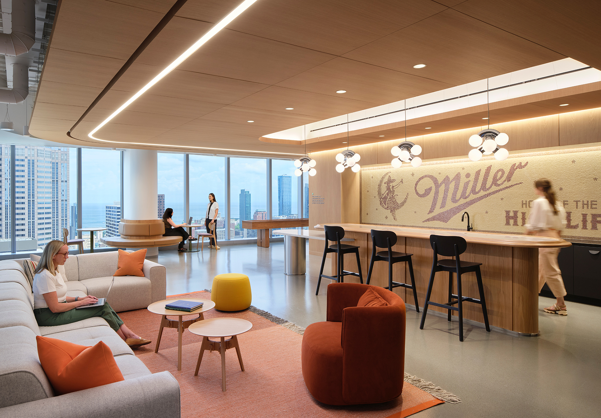 Modern office lounge with large windows, comfortable seating, a kitchenette with barstools, and “Miller” signage on the wall. People are working and conversing in a bright, open space overlooking the city.