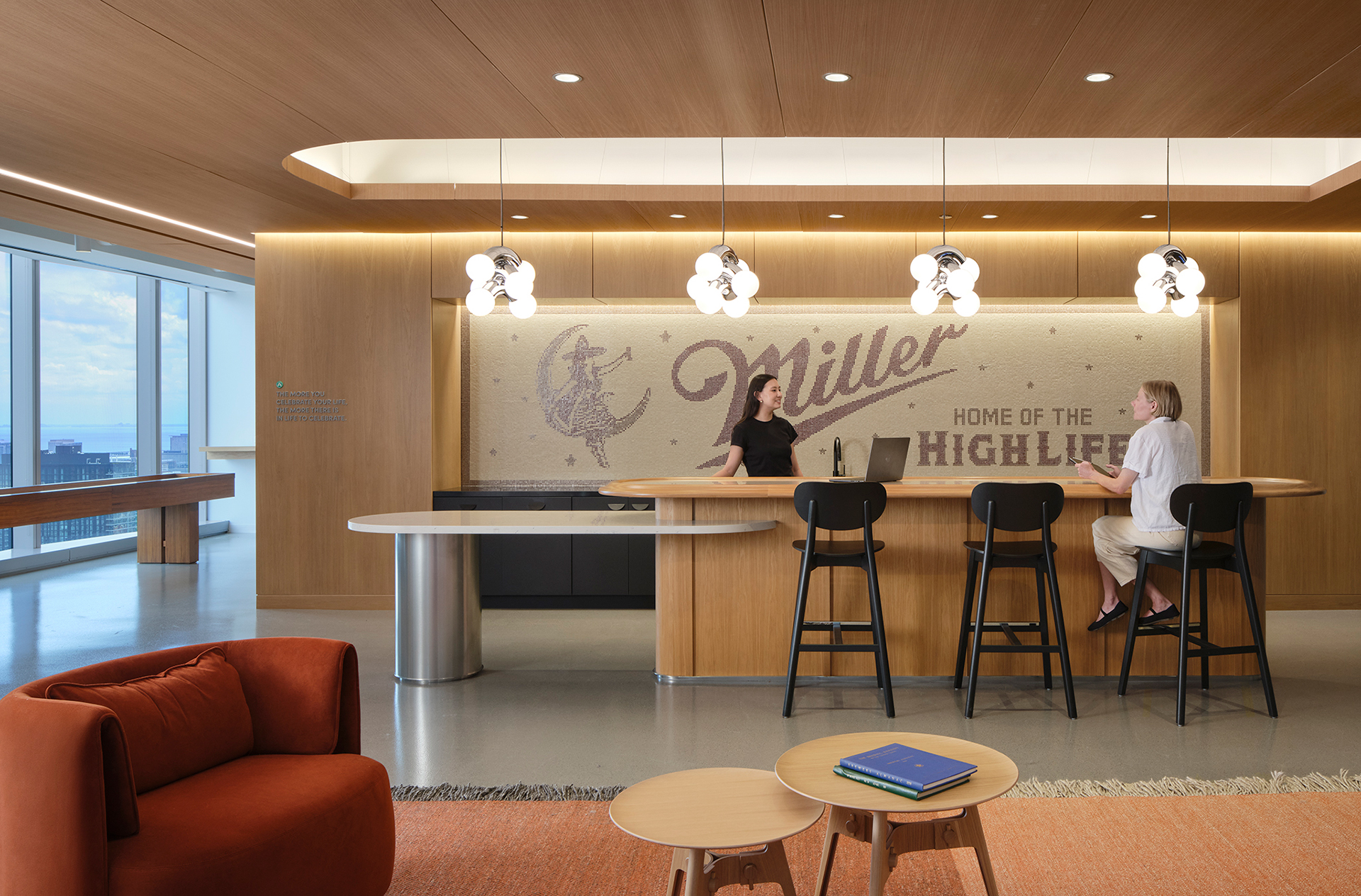 Modern office lobby with woodgrain acoustic felt accents, a round reception desk, two women talking, barstools, pendant lights, and a wall with “Miller Home of the High Life” and a moon graphic. Large windows reveal a city view.