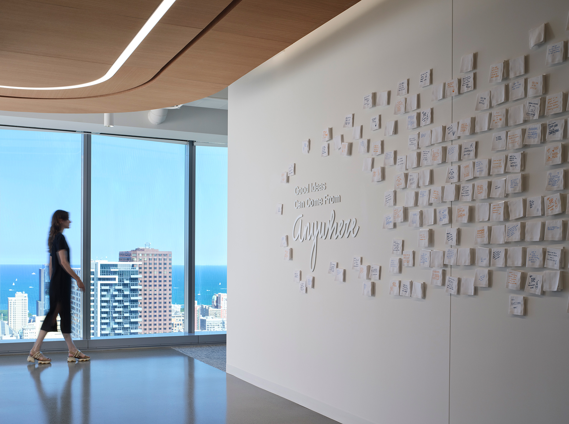 A woman walks through a modern office space with large windows overlooking a cityscape. On the wall are many small papers arranged around text that reads, “Good ideas can come from anywhere.”.