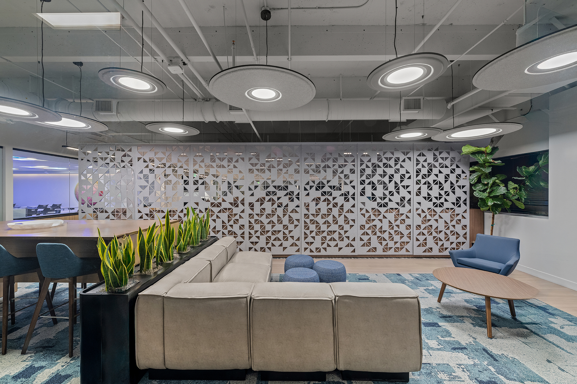 Modern office lounge with a gray sofa, a row of potted plants, blue chairs, round tables, geometric patterned acoustic felt wall screen, and circular ceiling lights. A TV and a large plant are in the corner on a blue-patterned rug.