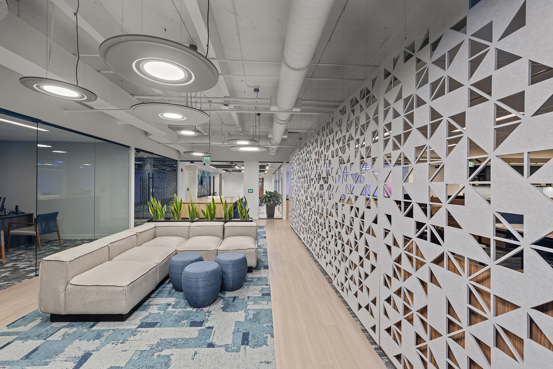 Modern office lobby with a beige sectional sofa, two blue textured ottomans, a geometric patterned acoustic felt wall screen, potted plants, and glass-walled offices, all under circular ceiling lights and exposed ductwork.