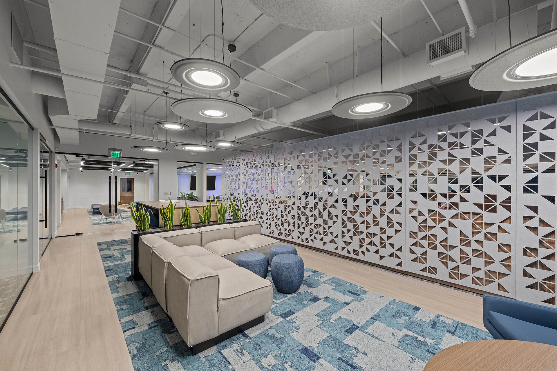 Modern office lounge with a beige sectional sofa, blue ottomans, patterned blue rug, decorative plant, and a geometric acoustic wall screens under circular ceiling lights. Glass walls and more seating are visible in the background.