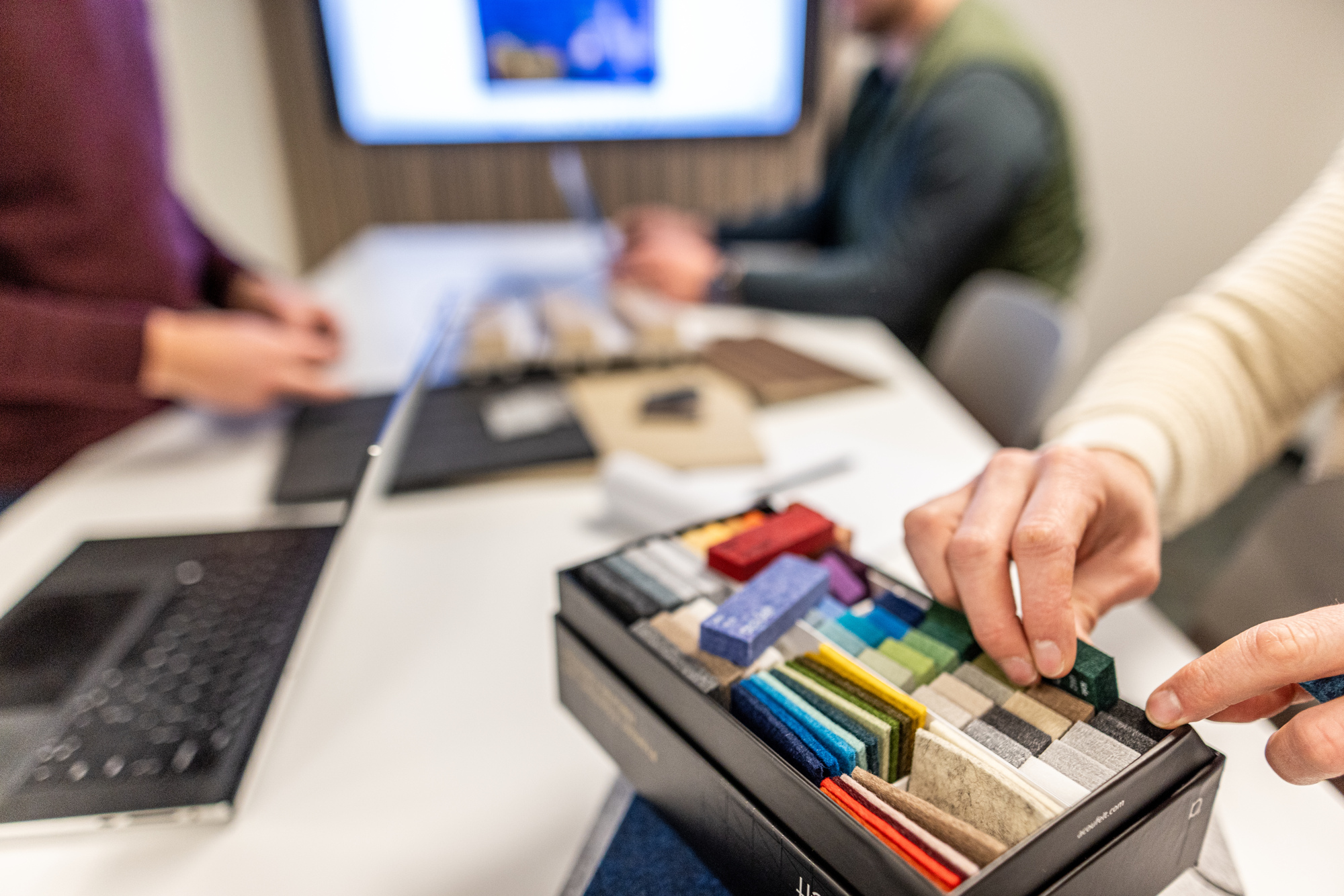 A person selecting a felt sample from a colorful sample box, with team members working in the blurred background of a studio,