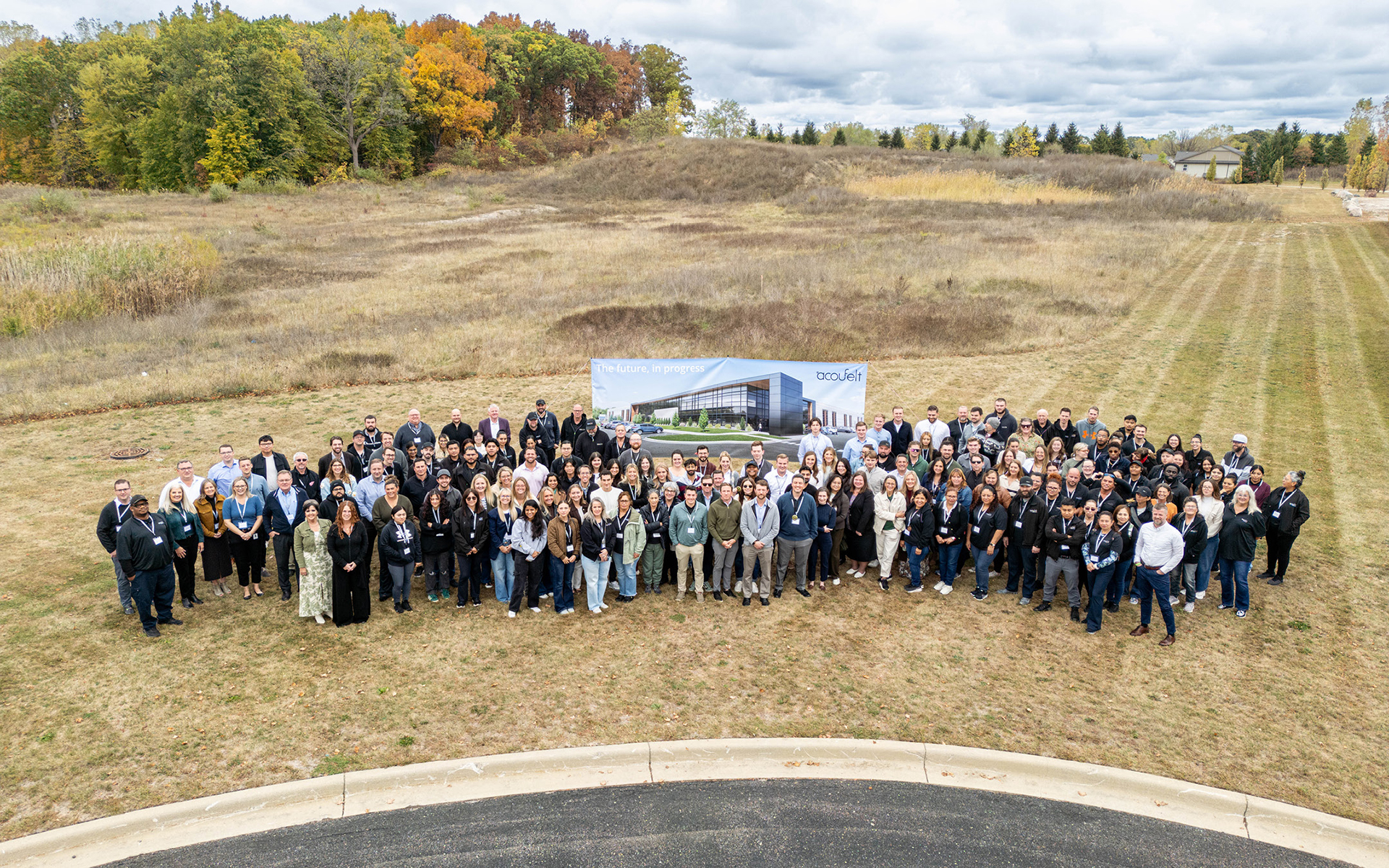 A large group of people pose for a group photo outdoors in front of a banner displaying a building rendering, with a grassy field and trees in the background under a cloudy sky.