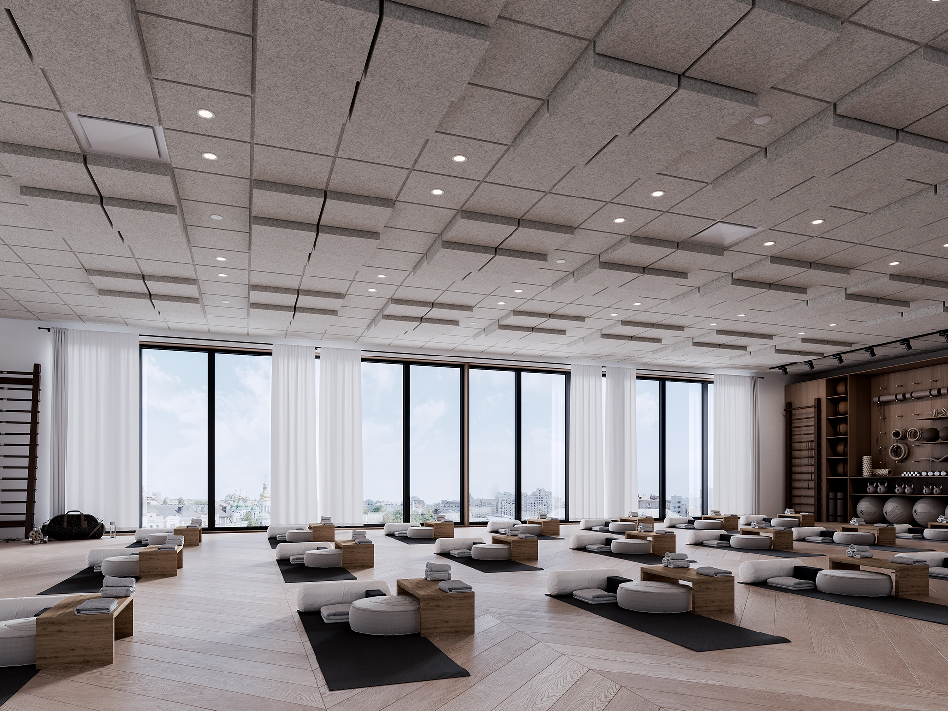 Spacious, modern yoga studio with large windows, acoustic felt ceiling, light wood floors, meditation cushions, yoga mats, and props arranged neatly in rows. Shelves with additional yoga equipment line the right wall.