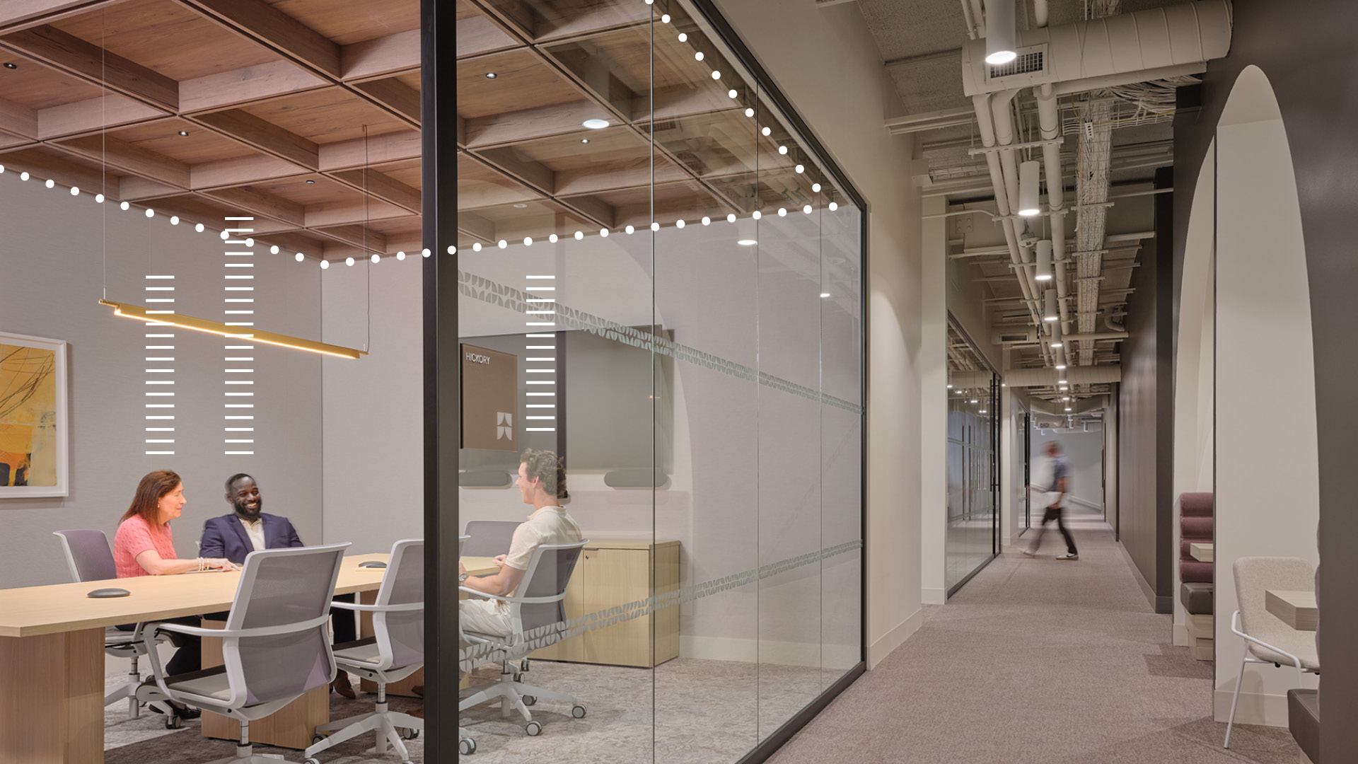 Three people sit around a table in a modern glass-walled conference room. A hallway runs alongside the room, with a blurred figure walking in the distance. The office space looks contemporary and well-lit.