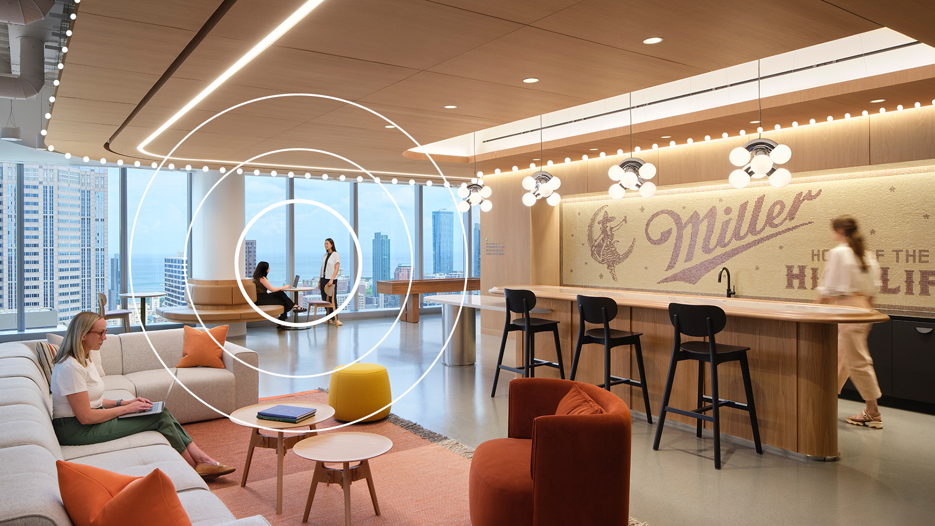Modern office lounge with large windows, comfortable seating, a kitchenette with barstools, and “Miller” signage on the wall. People are working and conversing in a bright, open space overlooking the city.